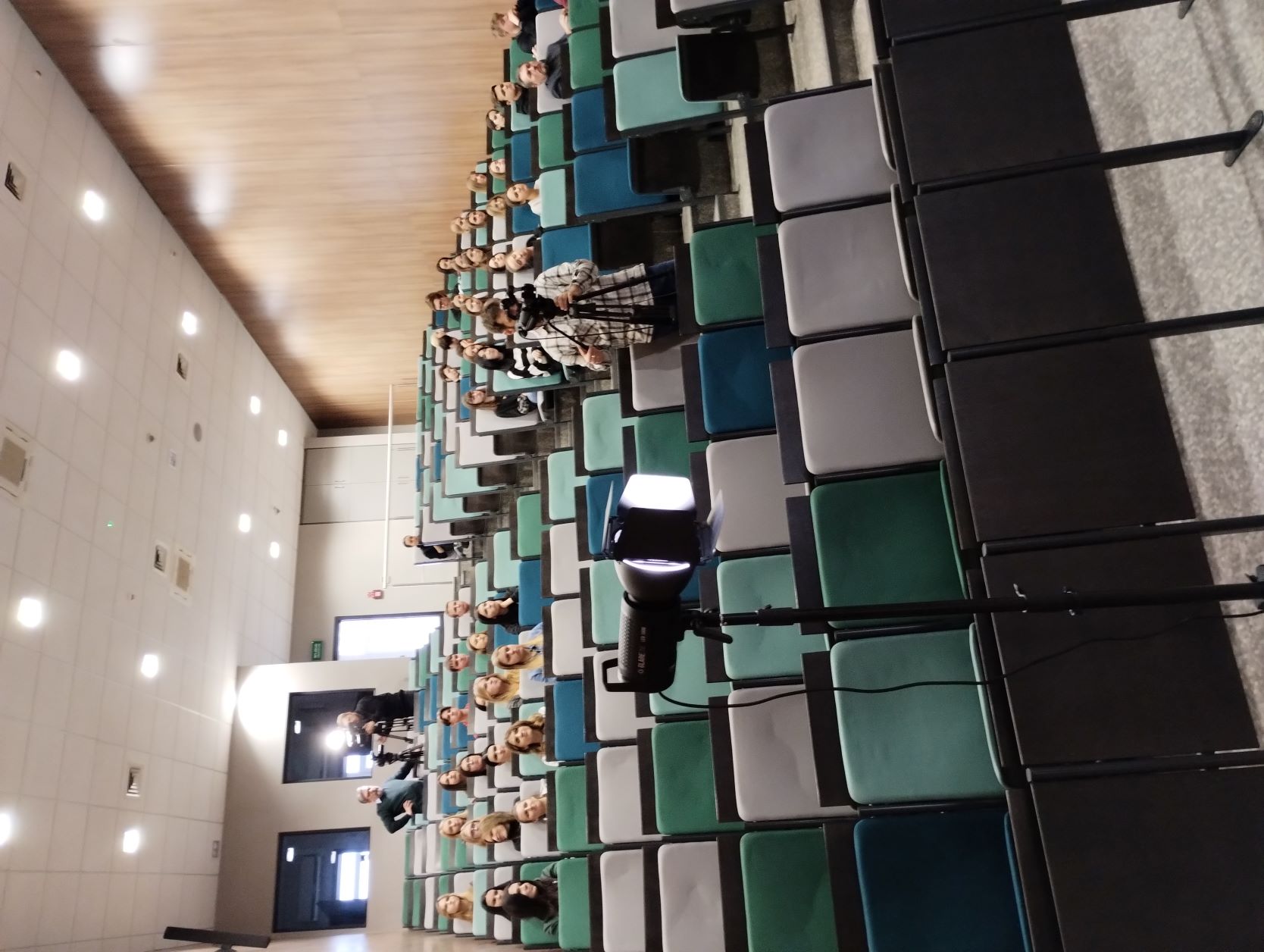 na aulia na krzesełkach siedzą uczestnicy wykładu. Widać na schodach także operatora kamery.