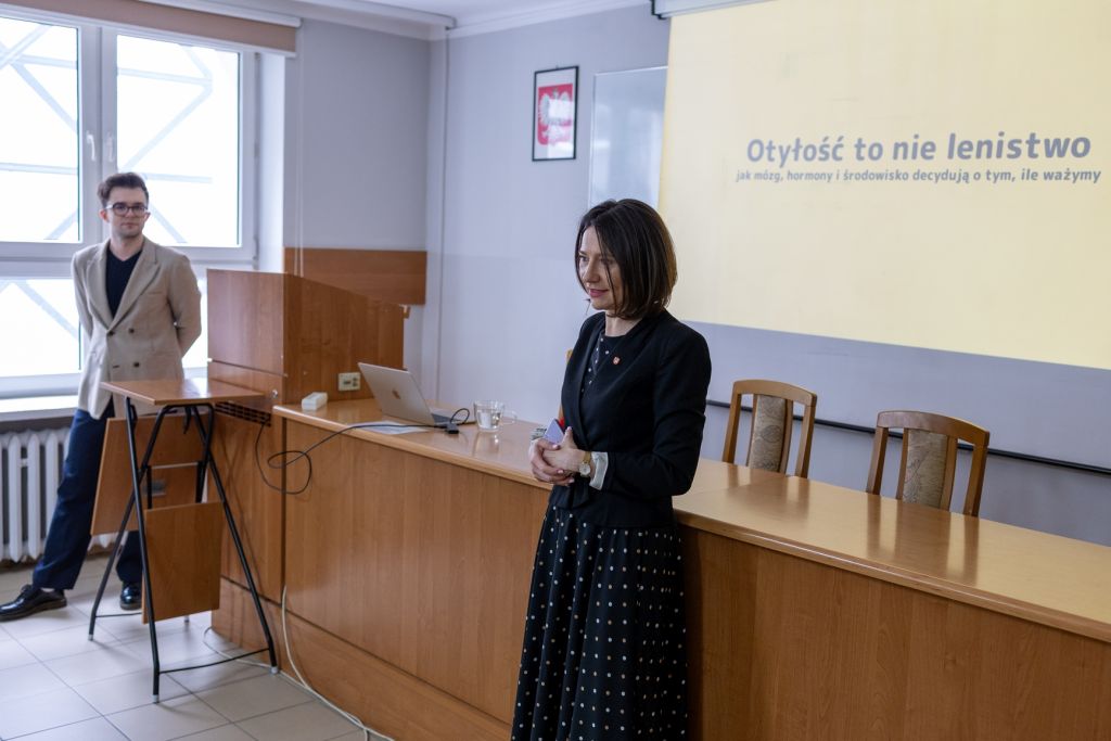 dr Kamila Sobaś i mgr Szymon Osowski stoją w sali wykładowej. Widać na ścianie Godło Polski oraz na ekranie projektora Otyłość to nie lenistwo, jak mózg, hormony i środowisko decydują o tym, ile ważymy