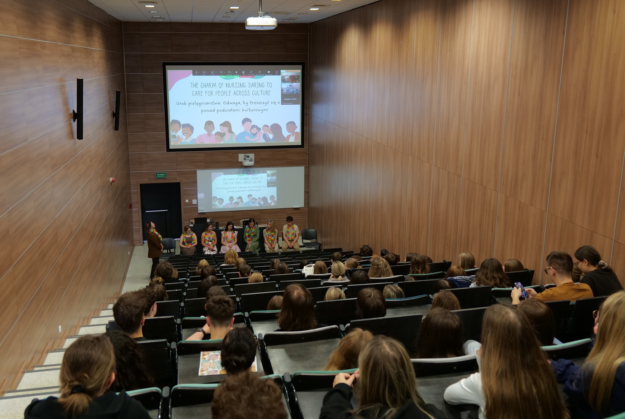 Sala wykładowa z drewnianymi panelami i rzędami stopniowanych siedzeń wypełnionych uczestnikami wydarzenia. Z przodu siedzi grupa osób biorących udział w prezentacji lub panelu dyskusyjnym, mających na szyjach kolorowe girlandy. Nad nimi wyświetlane są slajdy z ilustracjami przedstawiającymi różnorodne osoby, co podkreśla integracyjny i edukacyjny charakter spotkania.