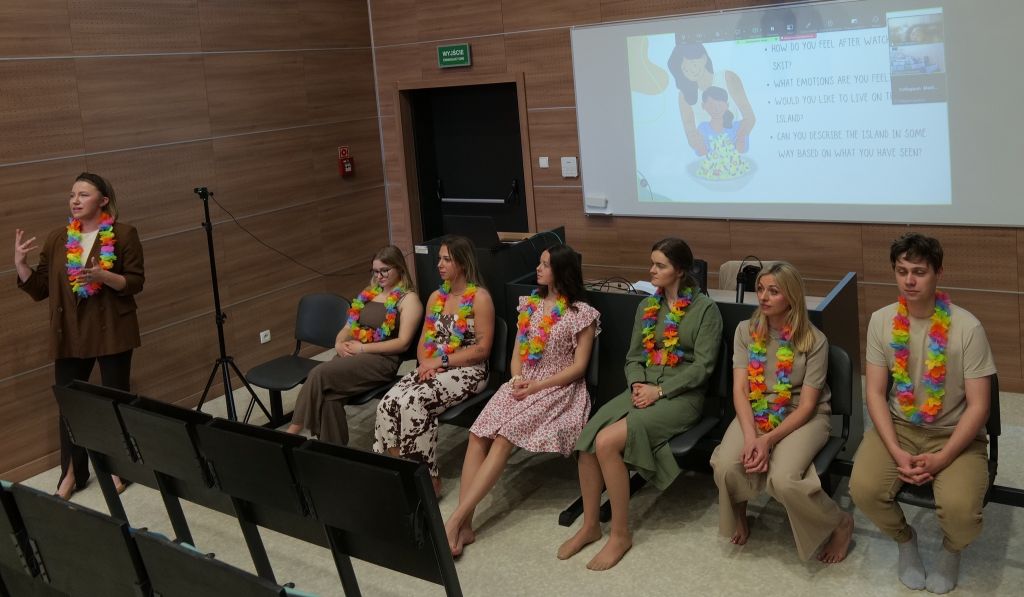 Sala dydaktyczna z uczestnikami siedzącymi w rzędzie podczas prezentacji lub warsztatu. Osoba prowadząca stoi z przodu i przemawia do grupy, a na ekranie wyświetlane są pytania oraz materiały prezentacyjne. Uczestnicy mają kolorowe girlandy na szyjach, a obok widoczna jest kamera na statywie, co wskazuje na rejestrowanie wydarzenia.