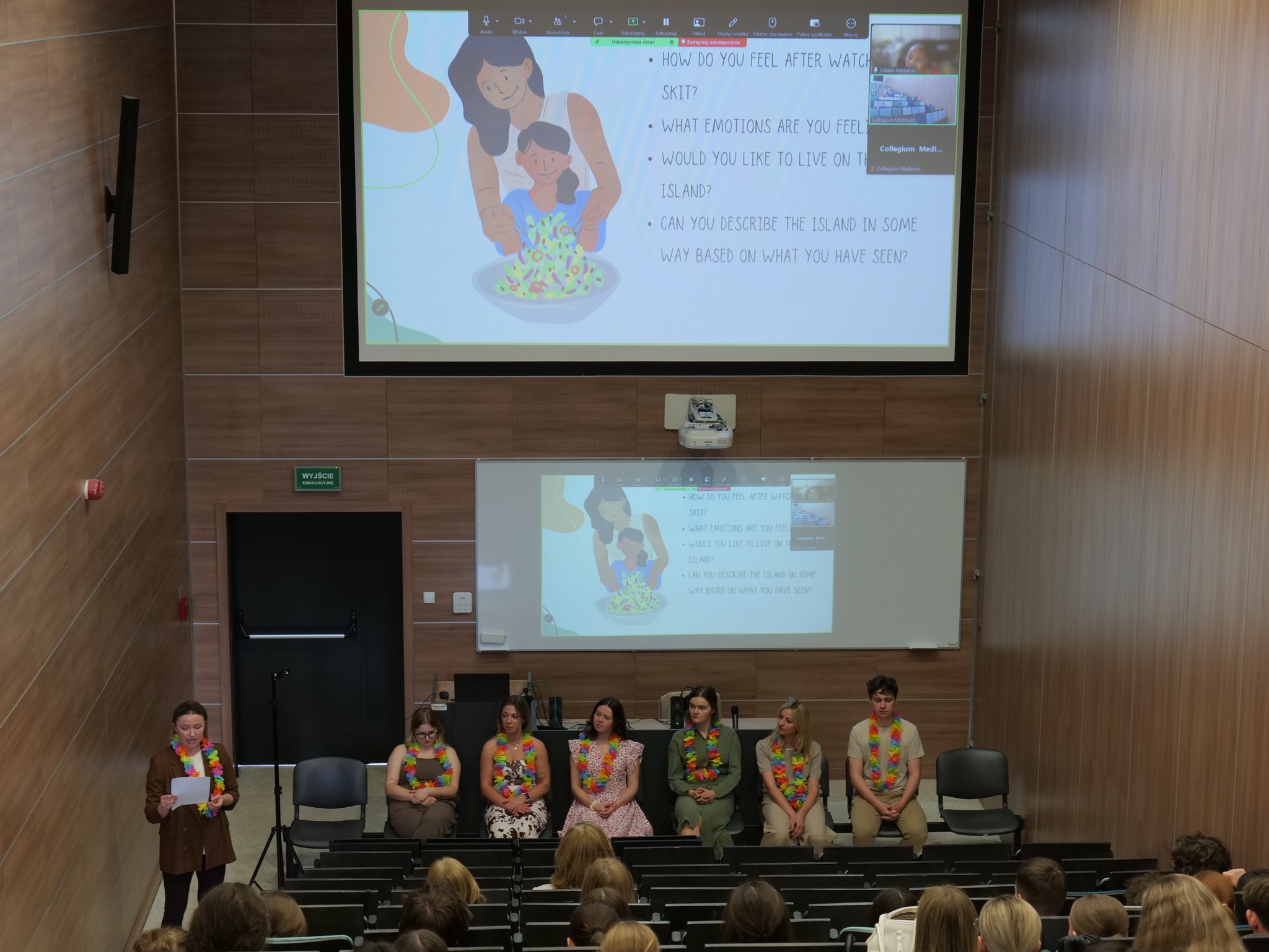 Sala wykładowa z drewnianymi panelami i dużym ekranem, przed którym odbywa się prezentacja lub panel dyskusyjny. Na scenie siedzi grupa uczestników z kolorowymi girlandami na szyjach, a jedna z osób prowadzi spotkanie, zwracając się do publiczności. Na ekranie widoczna jest ilustracja oraz pytania związane z omawianym scenariuszem, co podkreśla warsztatowy charakter wydarzenia.