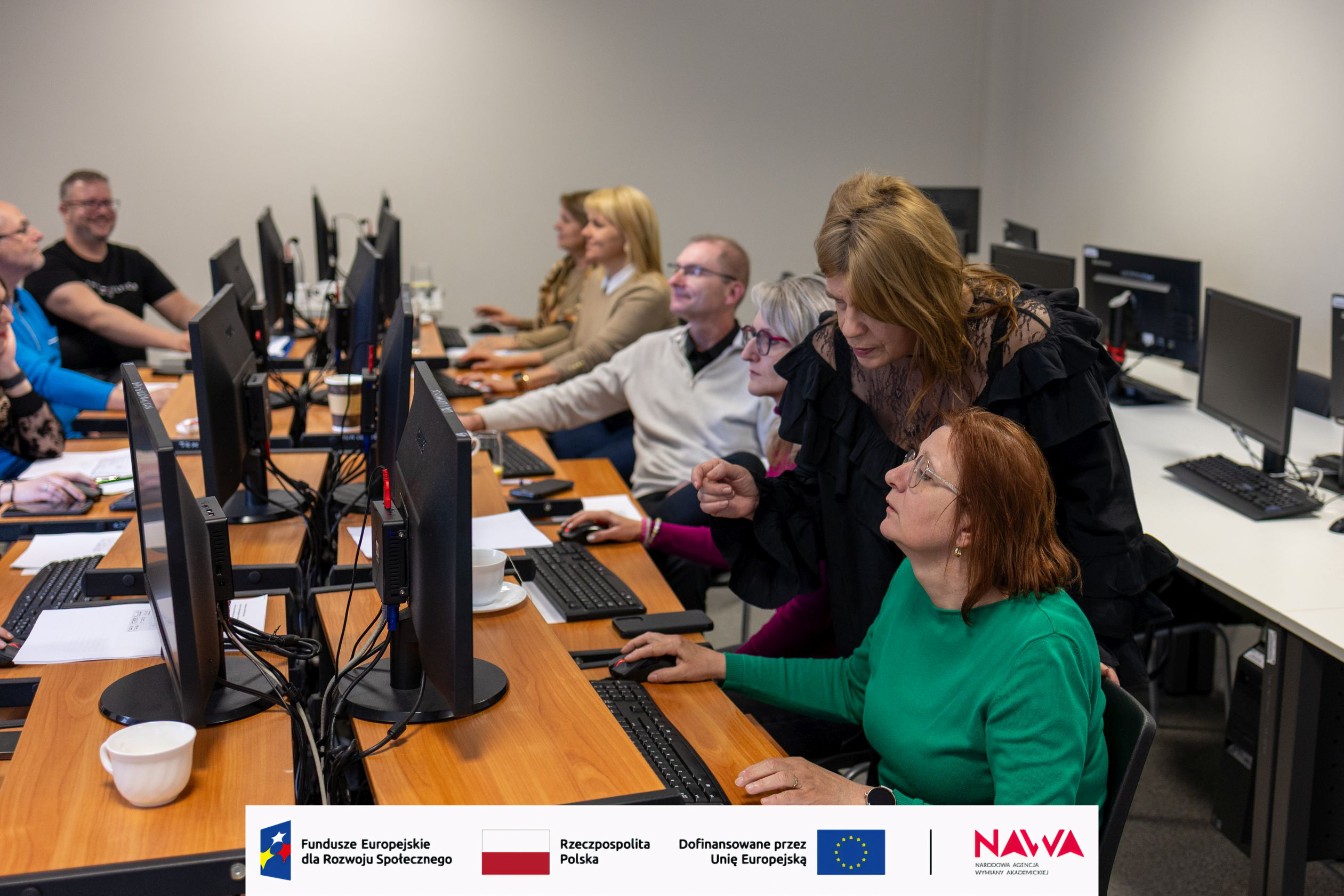 Sala komputerowa z uczestnikami pracującymi przy stanowiskach wyposażonych w monitory i klawiatury. Osoba prowadząca pomaga przy jednym z komputerów, wskazując elementy na ekranie i wspierając uczestników podczas zajęć. Na biurkach widoczne są notatki i materiały, a całość ma charakter szkoleniowy lub warsztatowy.