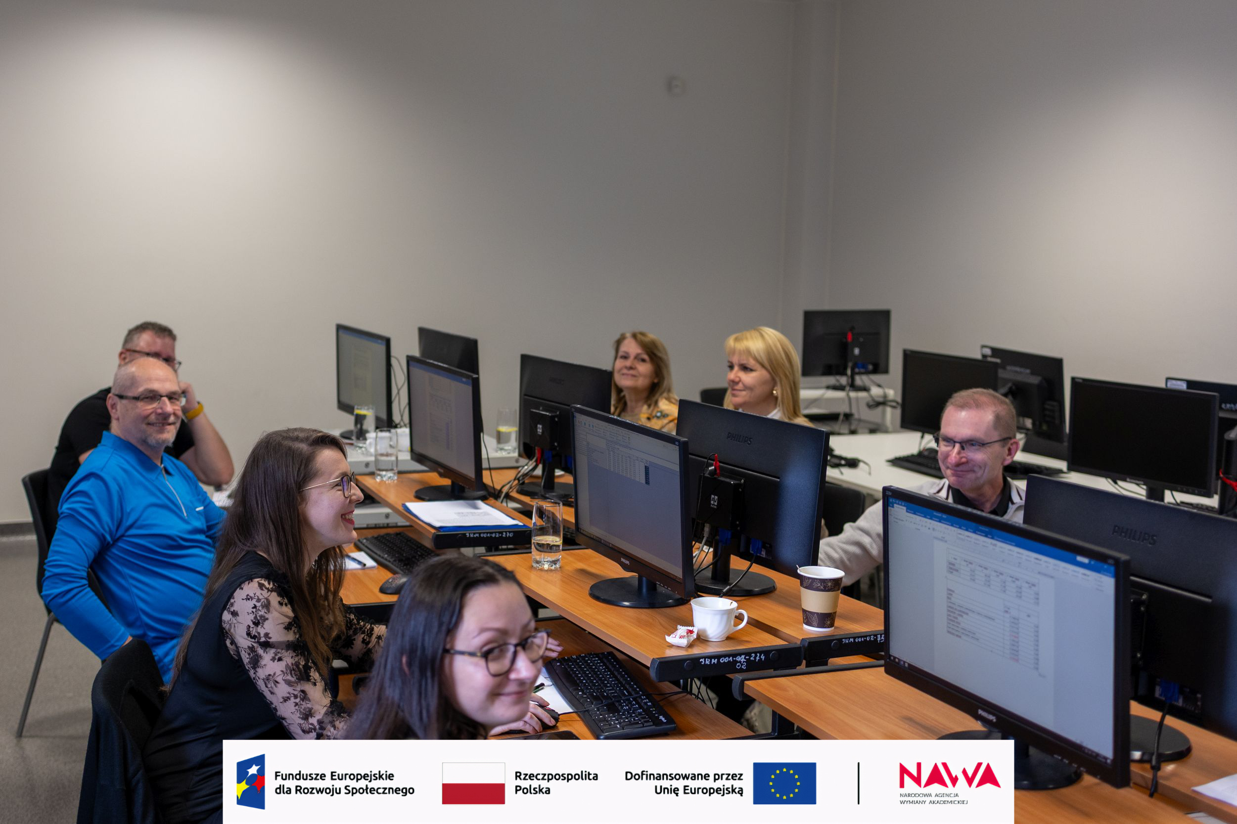 Sala komputerowa z uczestnikami siedzącymi przy stanowiskach i pracującymi na monitorach podczas szkolenia lub warsztatów. W pomieszczeniu ustawione są rzędy biurek z komputerami, a na stołach znajdują się kubki i notatki. Na ścianie widoczne są logotypy organizacji lub partnerów wydarzenia, co podkreśla profesjonalny charakter spotkania.