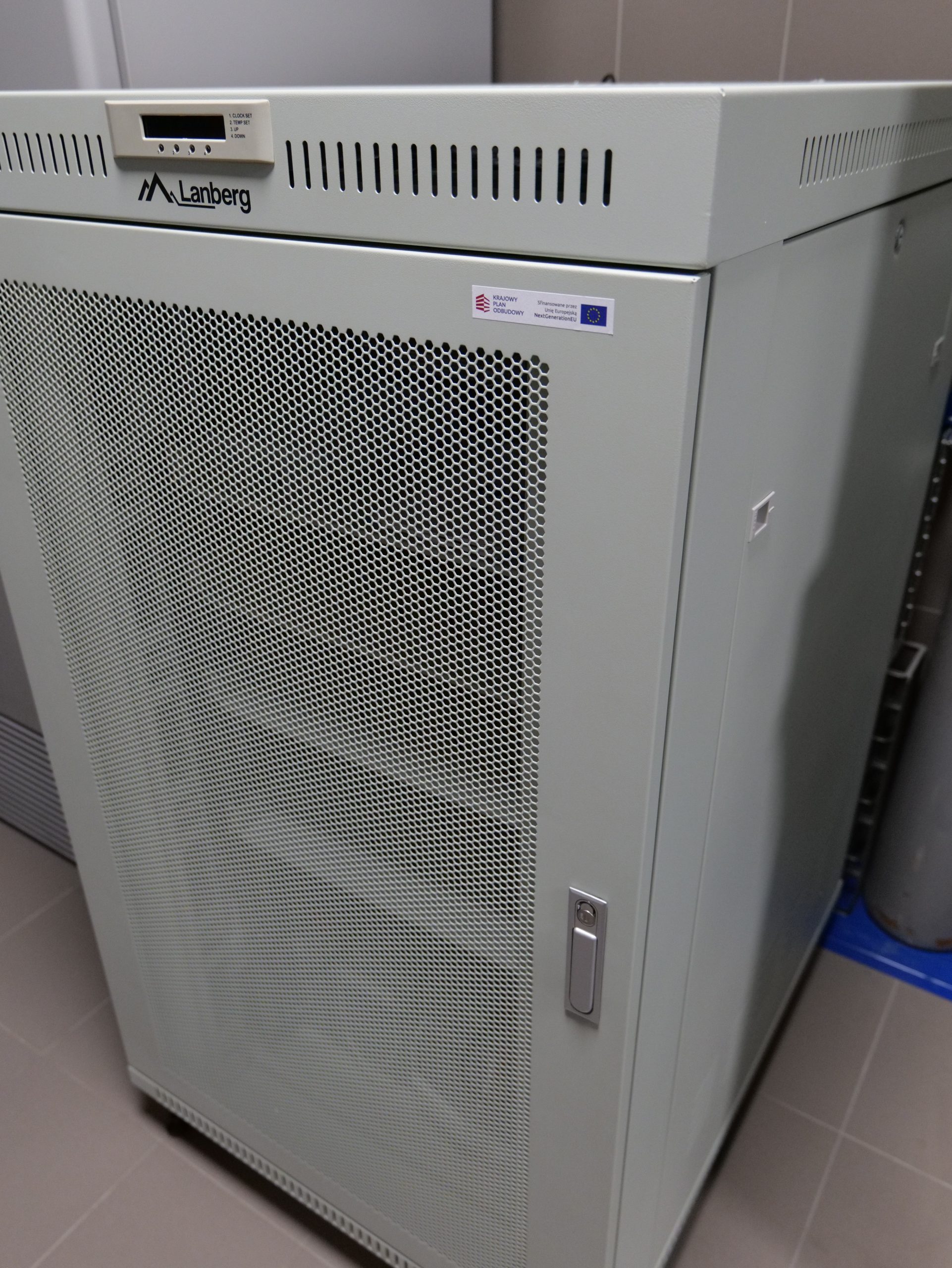 Szafa serwerowa przeznaczona do przechowywania i zabezpieczania urządzeń sieciowych oraz infrastruktury informatycznej. Konstrukcja wyposażona jest w perforowane drzwi zapewniające wentylację oraz panel monitorujący temperaturę pracy. Urządzenie znajduje się w pomieszczeniu technicznym o charakterze laboratoryjnym lub administracyjnym.