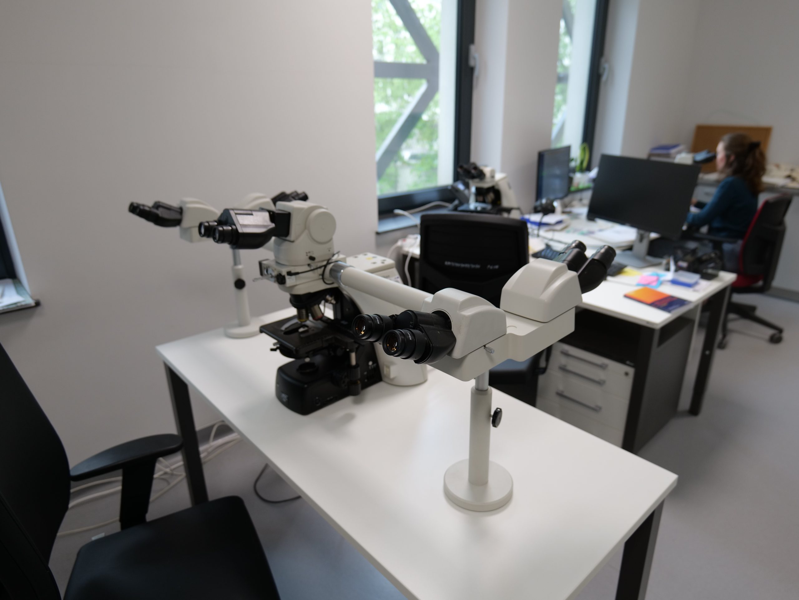Nowoczesne laboratorium wyposażone w zaawansowany mikroskop wielostanowiskowy umieszczony na stole roboczym. W tle znajduje się stanowisko komputerowe z monitorami, przy którym pracuje jedna z osób. Jasne wnętrze z dużymi oknami oraz specjalistycznym wyposażeniem podkreśla badawczy i dydaktyczny charakter pracowni.