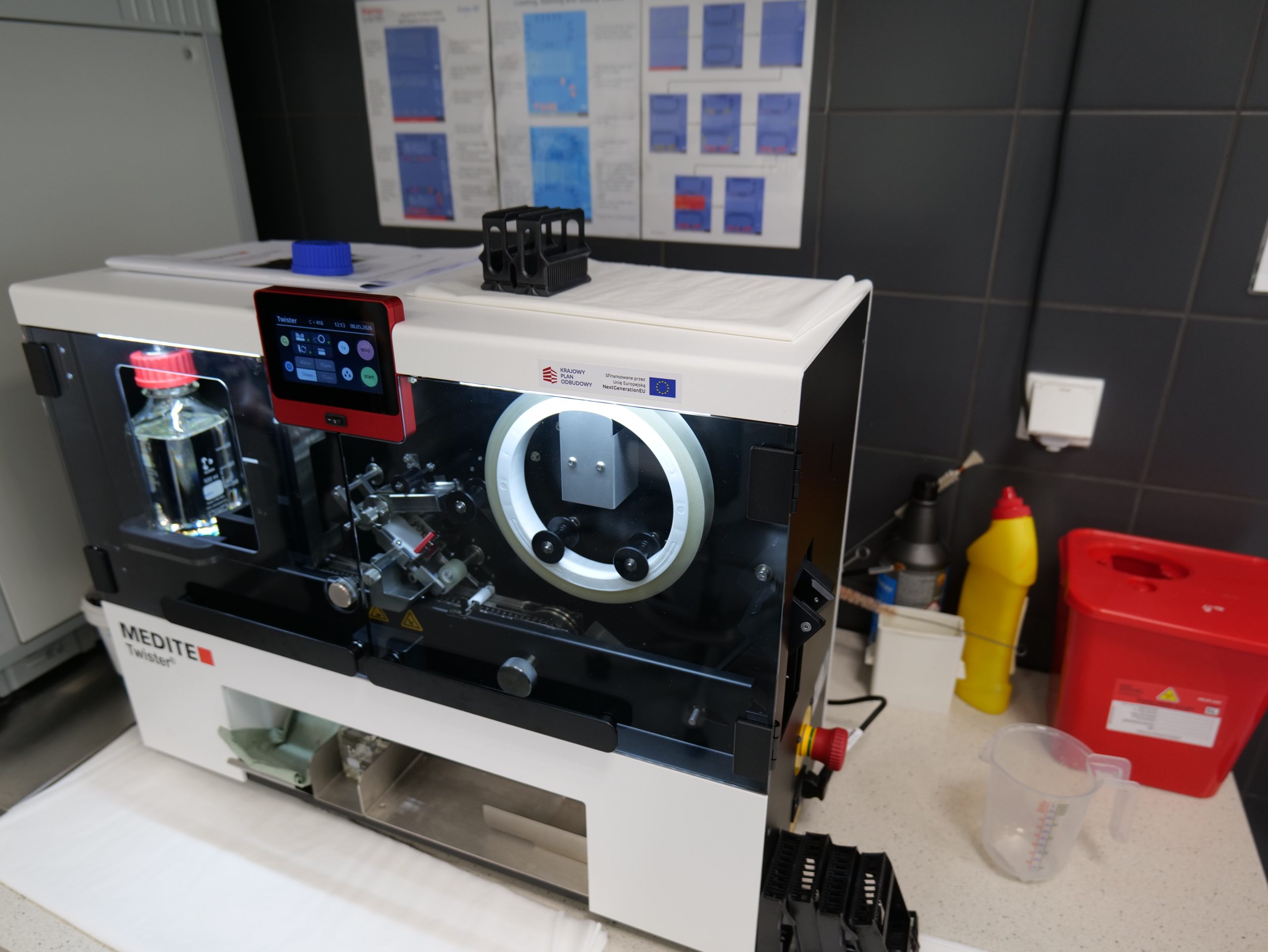Specjalistyczne urządzenie laboratoryjne „Medite Twister” ustawione na blacie roboczym w pracowni. Aparat wyposażony jest w ekran sterujący oraz zamkniętą komorę roboczą, w której widoczne są elementy mechaniczne i pojemniki na próbki. W otoczeniu znajdują się akcesoria laboratoryjne, pojemniki oraz materiały pomocnicze wykorzystywane podczas pracy laboratoryjnej.