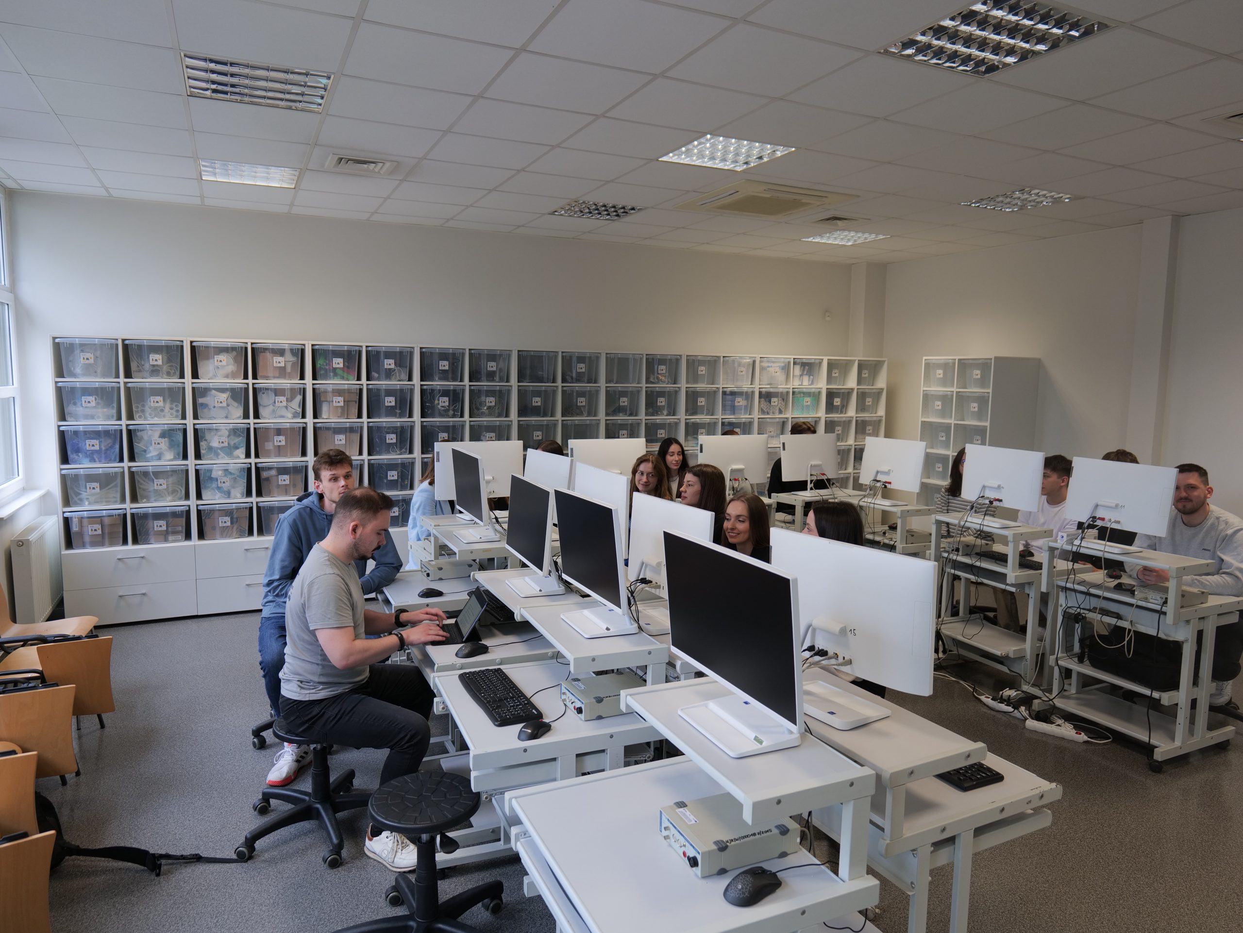 Nowoczesna pracownia komputerowa, w której uczestnicy pracują przy stanowiskach wyposażonych w komputery. Sala jest przestronna i dobrze oświetlona, z dużymi oknami zapewniającymi dostęp do światła dziennego. Wzdłuż ścian znajdują się regały z pojemnikami do przechowywania materiałów, a uczestnicy skupiają się na wykonywanych zadaniach w profesjonalnym i edukacyjnym środowisku.
