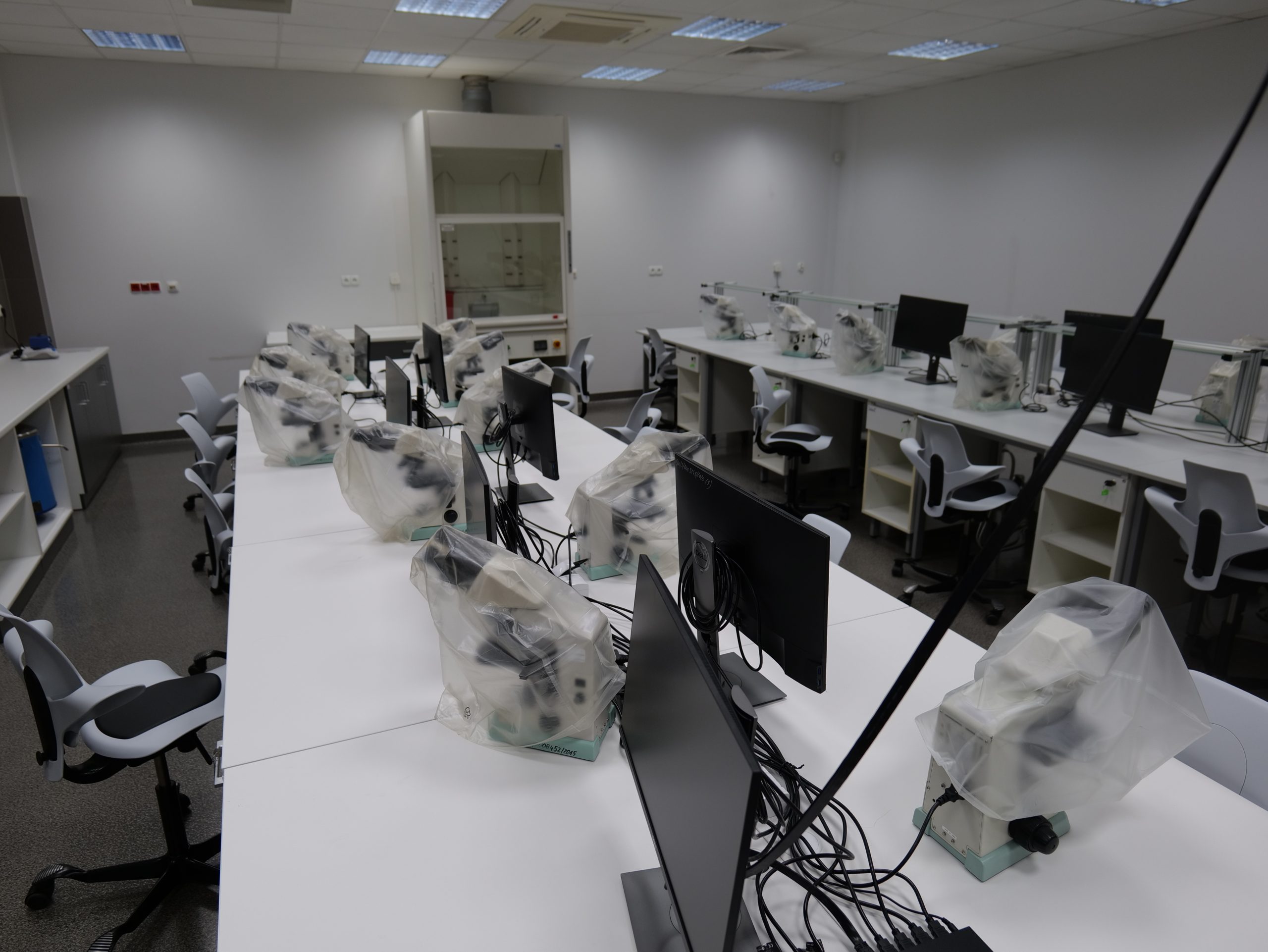Nowoczesna pracownia laboratoryjna wyposażona w stanowiska do pracy z mikroskopami i komputerami. Na długich stołach ustawiono mikroskopy połączone z monitorami, a przy stanowiskach znajdują się krzesła dla uczestników zajęć. W tle widoczny jest dygestorium laboratoryjne, a jasne oświetlenie i uporządkowane wyposażenie podkreślają dydaktyczny i badawczy charakter pomieszczenia.