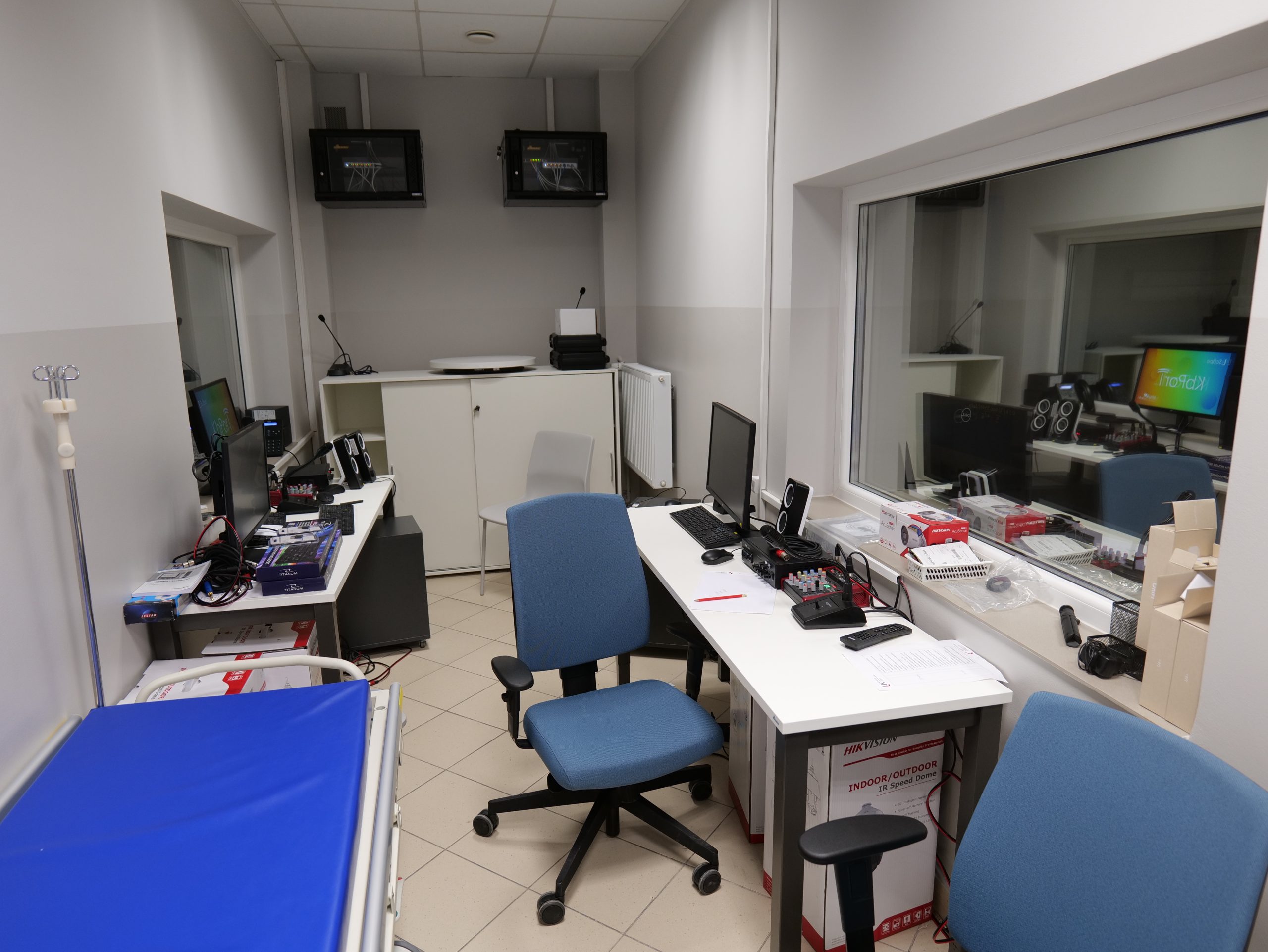 Pomieszczenie pełniące funkcję stanowiska obserwacyjnego lub sterowni w przestrzeni medycznej. Znajdują się w nim biurka z komputerami, monitorami oraz sprzętem technicznym służącym do nadzorowania zajęć lub badań. Przez dużą szybę można obserwować sąsiednie pomieszczenie, a dodatkowo widoczne są elementy wyposażenia medycznego, takie jak łóżko zabiegowe i stojak na kroplówki.