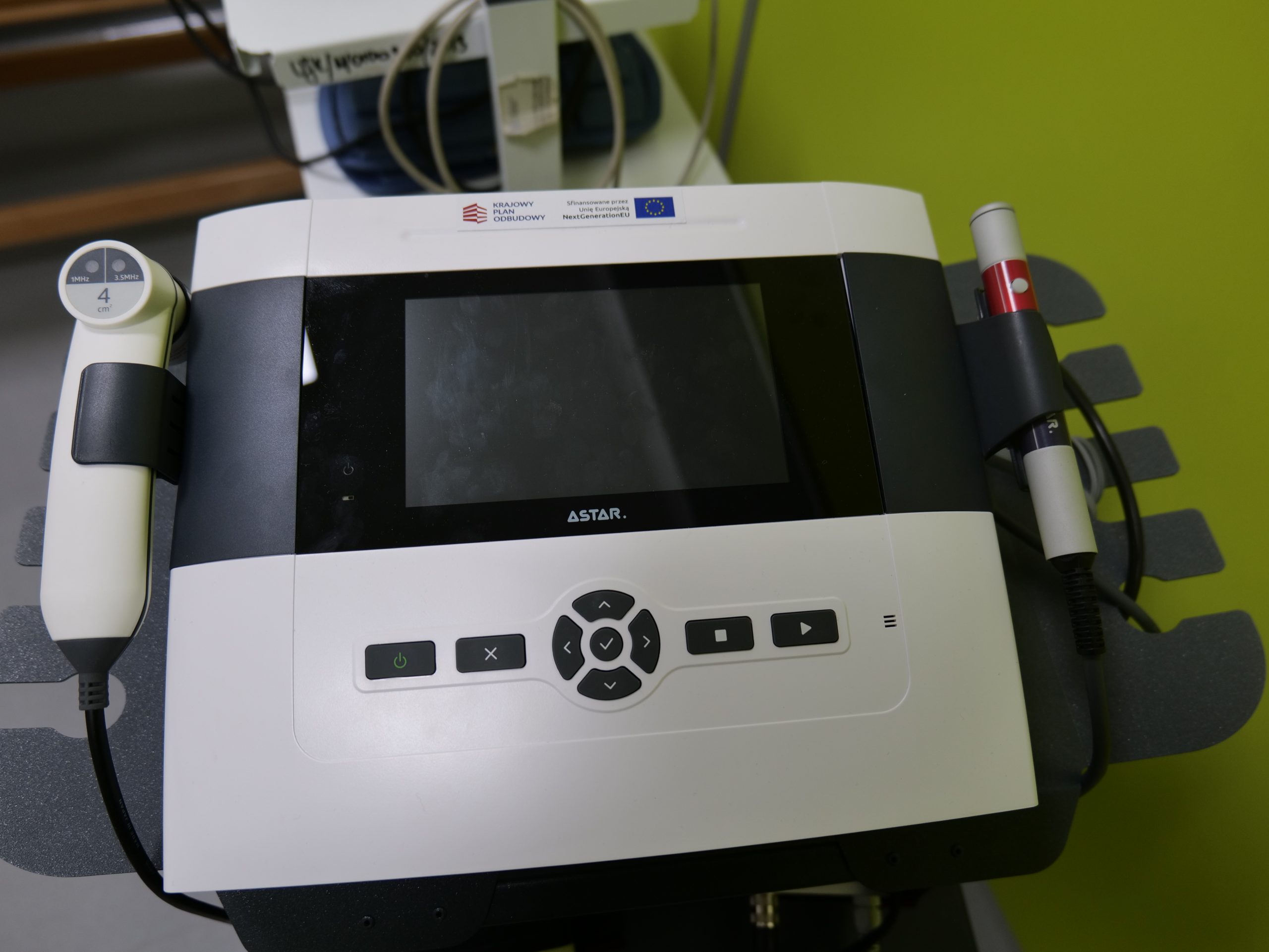 Urządzenie medyczne marki ASTAR przeznaczone do zabiegów fizykoterapeutycznych, wyposażone w ekran dotykowy oraz panel sterowania z przyciskami funkcyjnymi. Do aparatu podłączone są różne głowice zabiegowe służące do terapii, a całość umieszczona jest na mobilnym stojaku. W górnej części grafiki widoczne są logotypy partnerów lub instytucji wspierających projekt.