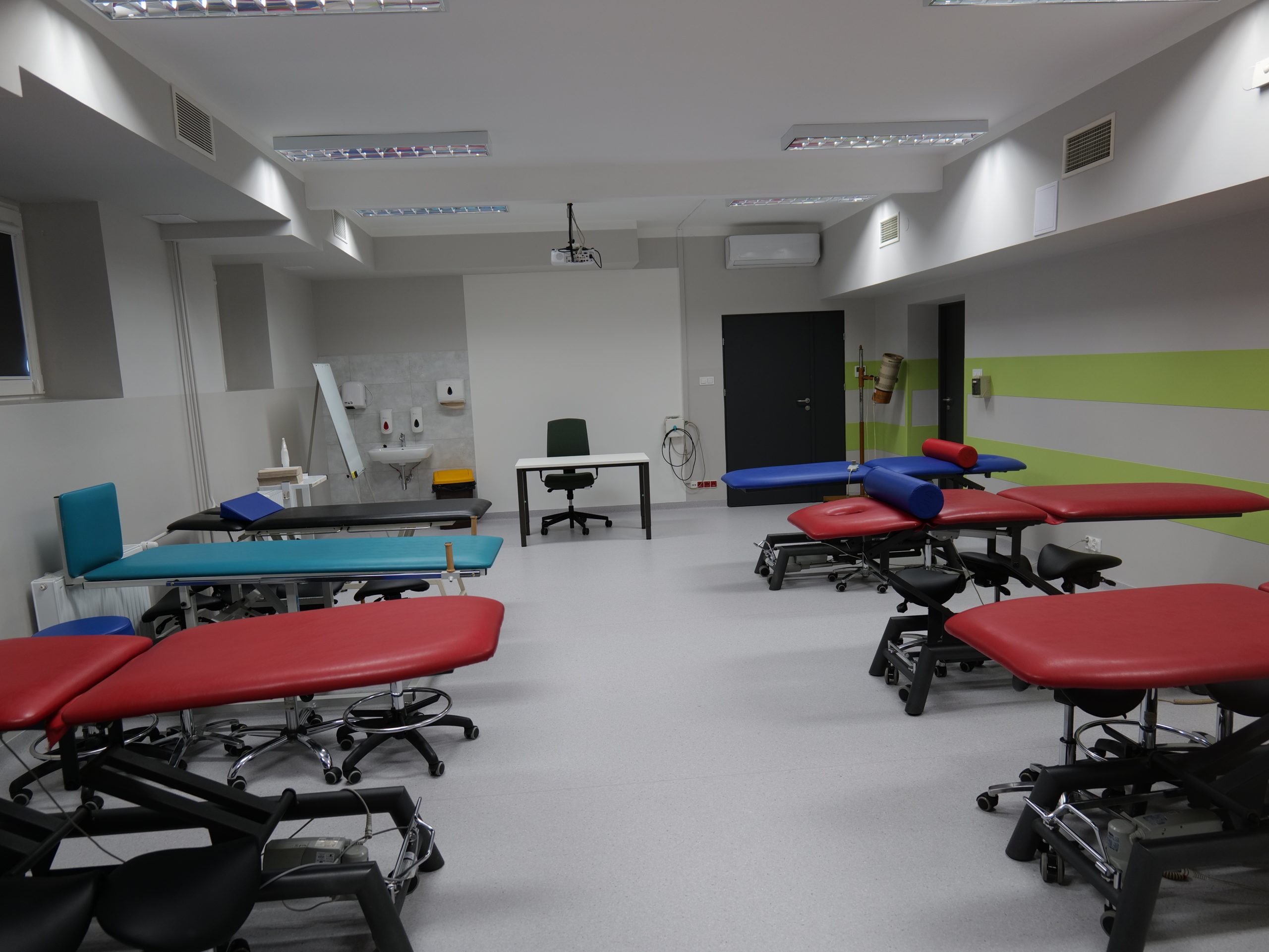 Przestronna sala dydaktyczna lub terapeutyczna wyposażona w kilka stołów rehabilitacyjnych i zabiegowych. Pomieszczenie jest dobrze oświetlone i przystosowane do prowadzenia zajęć praktycznych z zakresu fizjoterapii, rehabilitacji lub terapii manualnej. W sali znajdują się również stanowisko prowadzącego, umywalka oraz wyposażenie zapewniające odpowiednie warunki do nauki i ćwiczeń praktycznych.