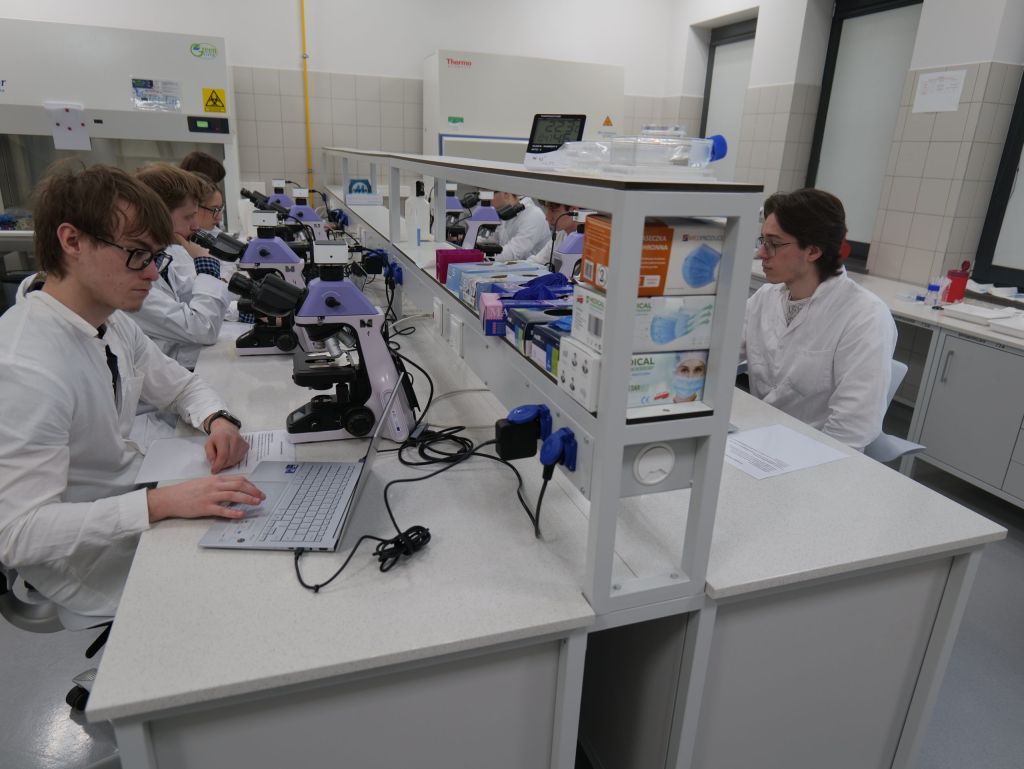 Studenci pracują w nowoczesnym laboratorium przy wspólnym stanowisku wyposażonym w mikroskopy i sprzęt komputerowy. Uczestnicy zajęć, ubrani w fartuchy laboratoryjne, wykonują obserwacje preparatów oraz analizują wyniki badań. Pracownia jest dobrze wyposażona i utrzymana w standardzie typowym dla zajęć dydaktycznych oraz pracy badawczej.