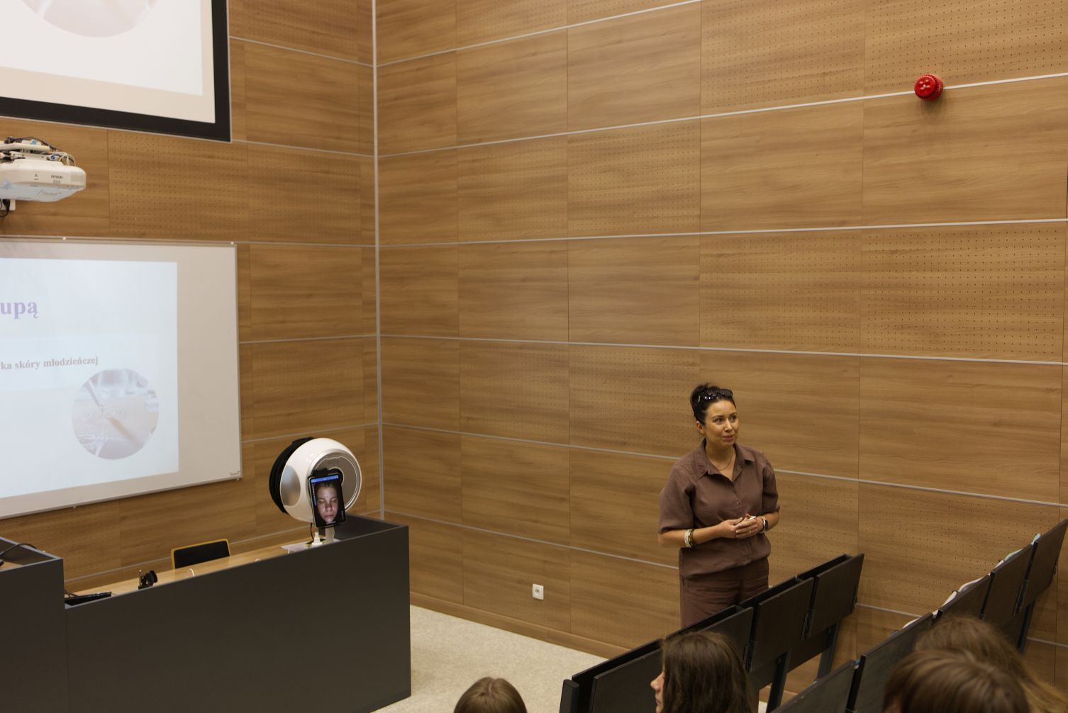 Nowoczesna aula lub sala wykładowa została wyposażona w sprzęt multimedialny oraz specjalistyczne urządzenia wykorzystywane podczas prezentacji i pokazów praktycznych. W pobliżu mównicy znajduje się aparat diagnostyczny z ekranem prezentującym wyniki badania lub obraz uczestnika. Drewniane wykończenie wnętrza oraz amfiteatralny układ miejsc podkreślają akademicki charakter wydarzenia.