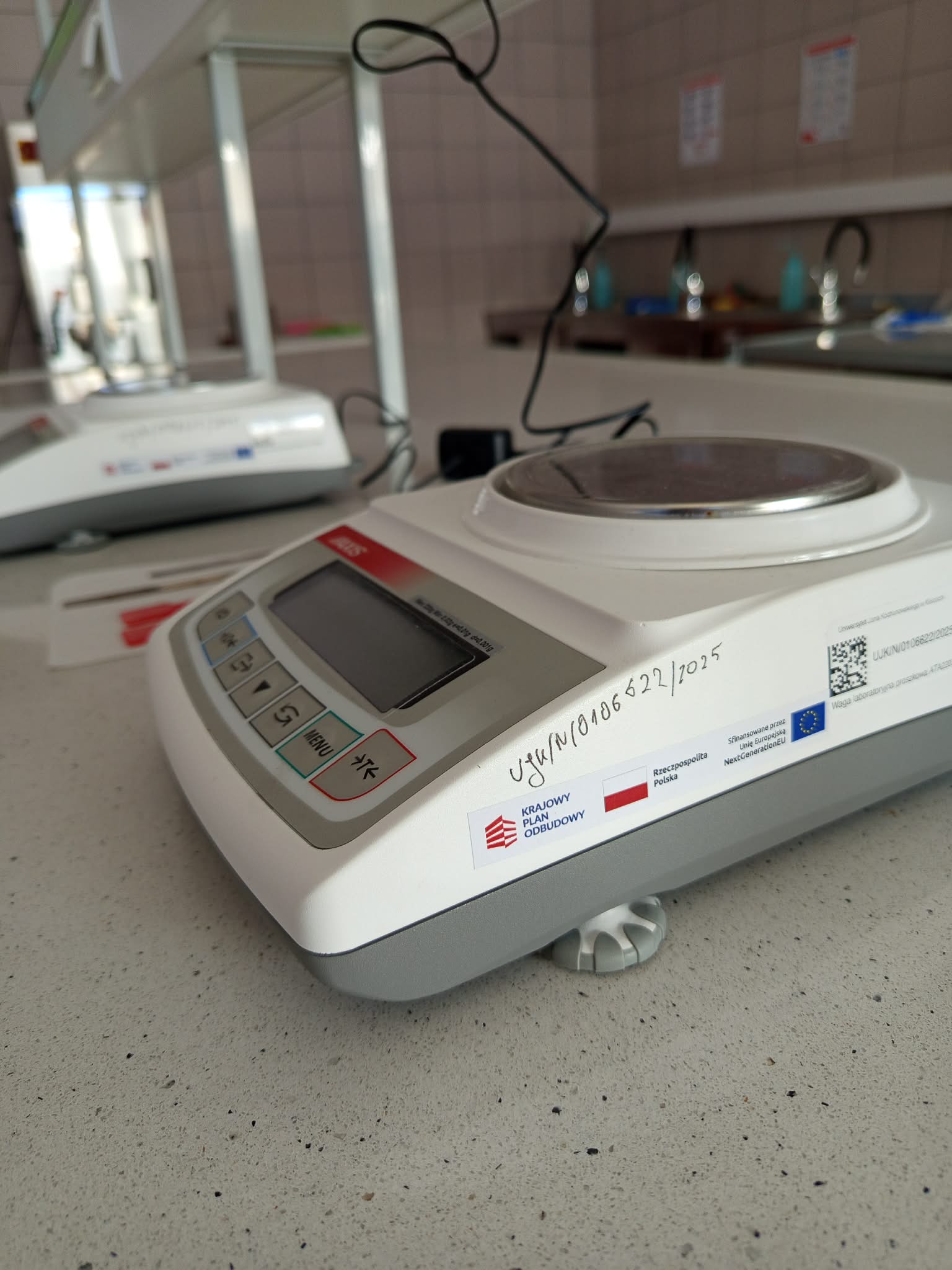 Zdjęcie przedstawia laboratoryjne urządzenie grzewcze lub mieszające z okrągłą płytą ze stali nierdzewnej, wyświetlaczem i przyciskami sterującymi. Na obudowie widoczny jest odręczny numer identyfikacyjny oraz etykiety dotyczące finansowania ze środków polskich i Unii Europejskiej z kodem QR. W tle znajdują się dodatkowe urządzenia laboratoryjne, zlew, butelki oraz wyposażenie pomieszczenia.