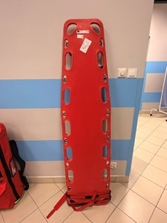 Zdjęcie przedstawia czerwoną deskę ortopedyczną do transportu poszkodowanych, ustawioną pionowo przy ścianie. Deska posiada liczne uchwyty, otwory mocujące oraz pasy zabezpieczające. W tle widoczne są torba ratownicza, gniazda elektryczne i fragment wyposażenia pomieszczenia.