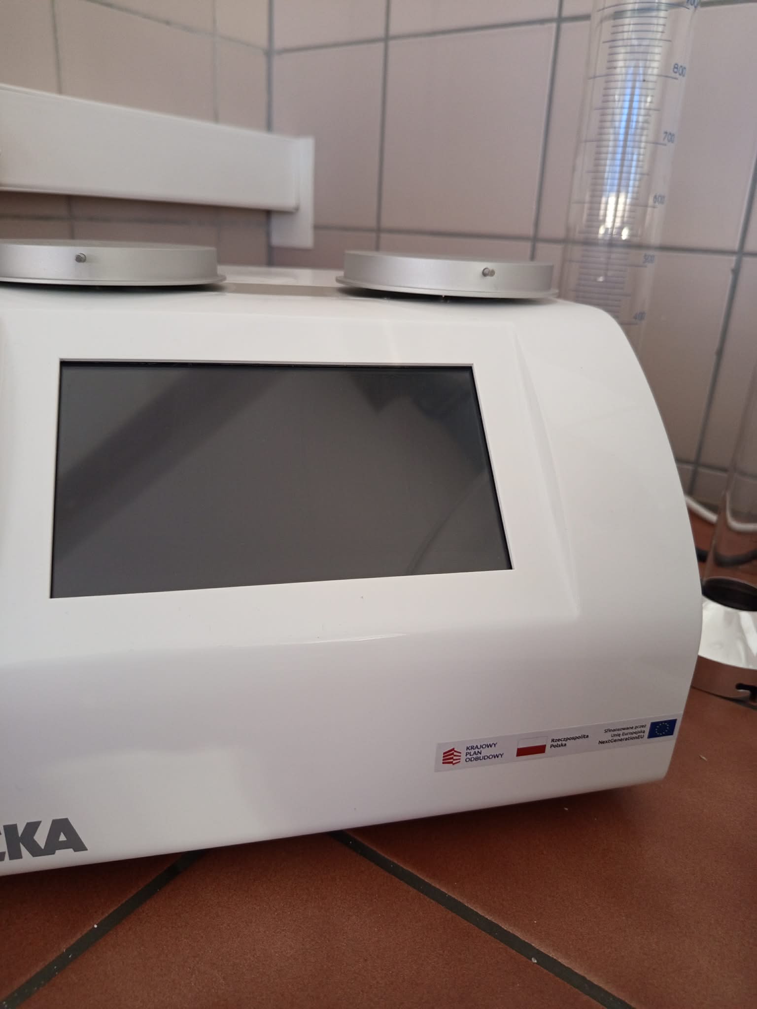 Zdjęcie przedstawia białe urządzenie laboratoryjne IKA z dużym, nieaktywnym wyświetlaczem oraz dwoma pokrętłami sterującymi. Obok znajduje się przezroczysty cylinder miarowy z oznaczeniami objętości. Na obudowie widoczna jest etykieta dotycząca finansowania ze środków polskich i Unii Europejskiej.