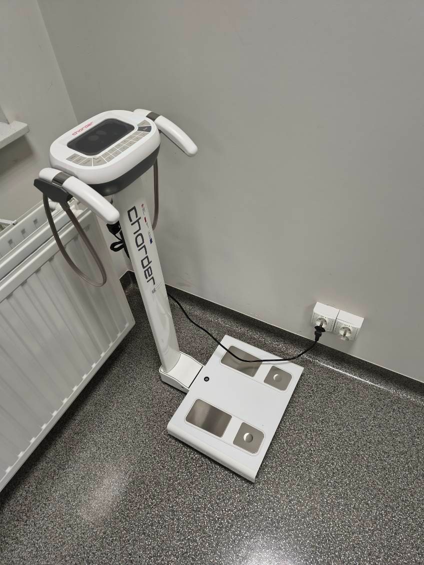 Zdjęcie przedstawia urządzenie do analizy składu ciała wyposażone w panel sterowania, uchwyty oraz platformę pomiarową. Na podstawie znajdują się metalowe elektrody przeznaczone do kontaktu ze stopami. Urządzenie jest podłączone do zasilania i ustawione przy ścianie w pomieszczeniu diagnostycznym.