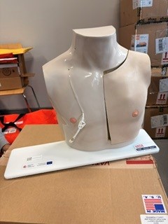 Zdjęcie przedstawia biały, anatomiczny manekin tułowia umieszczony na podstawie ekspozycyjnej. Na jego powierzchni widoczne są oznaczenia punktów pomiarowych oraz linie pomocnicze. W tle znajdują się regały, kartony i inne wyposażenie przestrzeni magazynowej lub warsztatowej.