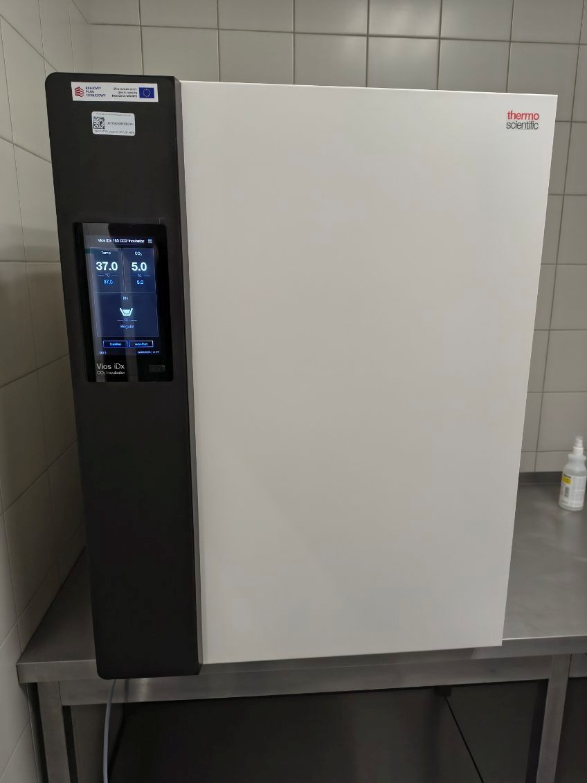 Zdjęcie przedstawia inkubator CO₂ Thermo Scientific Vios iDx ustawiony na laboratoryjnym blacie ze stali nierdzewnej. Na panelu sterowania widoczny jest ekran pokazujący temperaturę 37,0°C oraz poziom CO₂ wynoszący 5,0%. Wokół urządzenia widoczne są oznaczenia, naklejka z kodem QR oraz dodatkowe wyposażenie laboratoryjne.