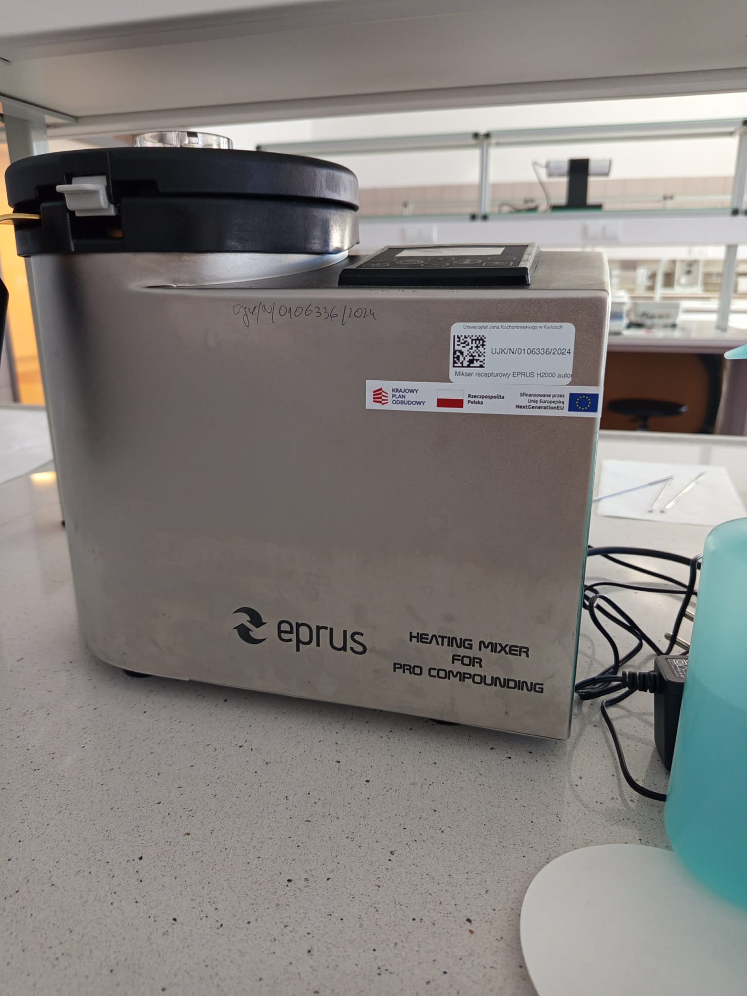 Zdjęcie przedstawia laboratoryjny mieszalnik z funkcją podgrzewania EPRUS, ustawiony na blacie roboczym. Urządzenie posiada komorę mieszającą, pokrywę oraz panel sterowania, a na froncie widoczne jest oznaczenie „HEATING MIXER FOR PRO COMPOUNDING”. Na obudowie znajdują się również etykiety identyfikacyjne i informacje dotyczące finansowania, a w tle widoczne jest dodatkowe wyposażenie laboratoryjne.