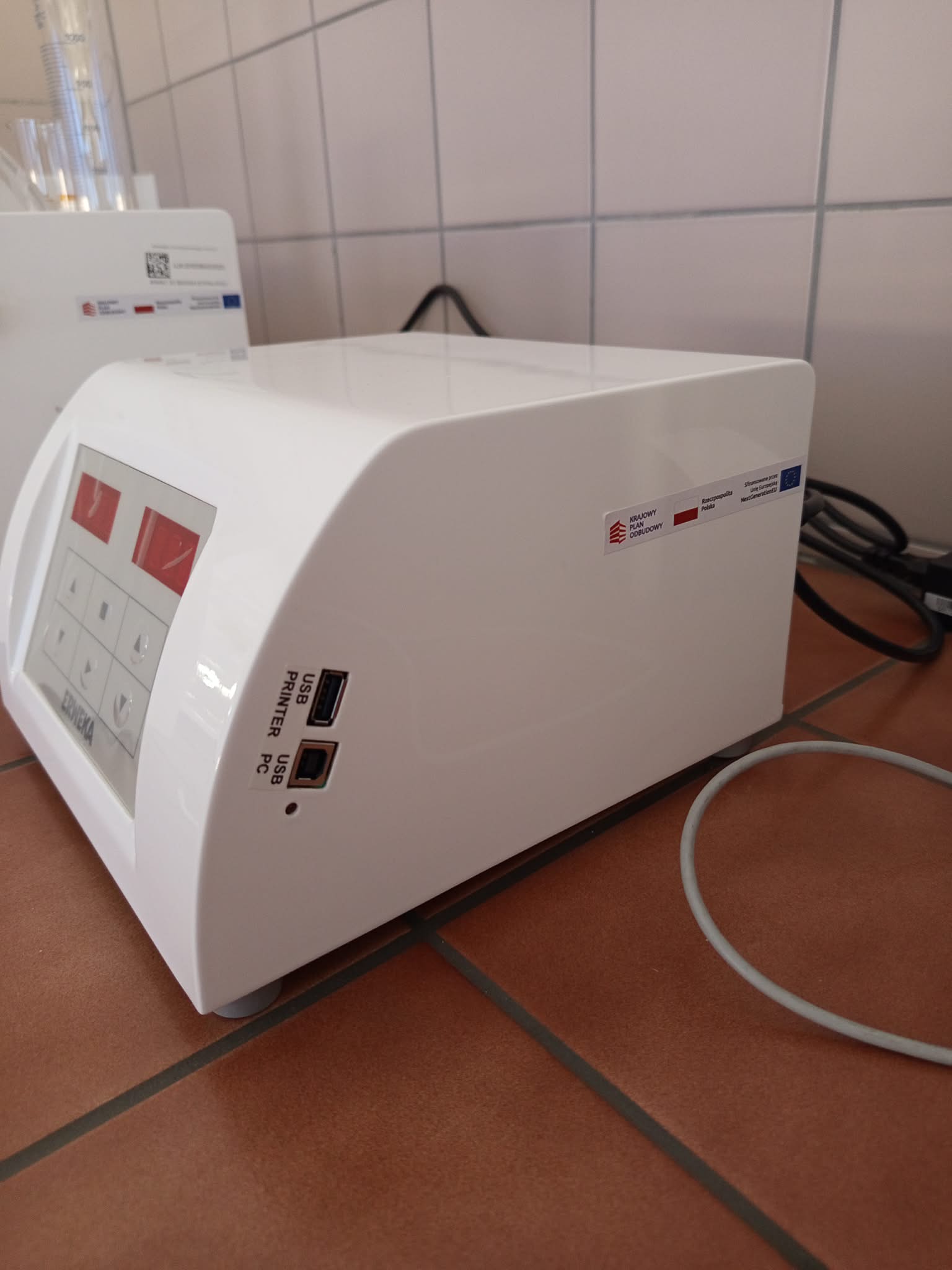 Zdjęcie przedstawia białą drukarkę laboratoryjną lub medyczną z panelem sterowania wyposażonym w przyciski. Na froncie znajdują się porty USB przeznaczone do podłączenia drukarki i komputera, a na obudowie widoczne są etykiety dotyczące finansowania ze środków polskich i Unii Europejskiej. Do urządzenia podłączone są przewody, a w tle widoczne są dodatkowe pojemniki laboratoryjne.