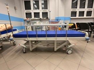 Zdjęcie przedstawia łóżko transportowe na kółkach znajdujące się w sali klinicznej. Łóżko wyposażone jest w niebieski materac, podnoszone barierki, regulowane podpory oraz duże koła jezdne. Wokół widoczne są urządzenia i przewody medyczne, a także dodatkowe łóżko, krzesła i wyposażenie sali.