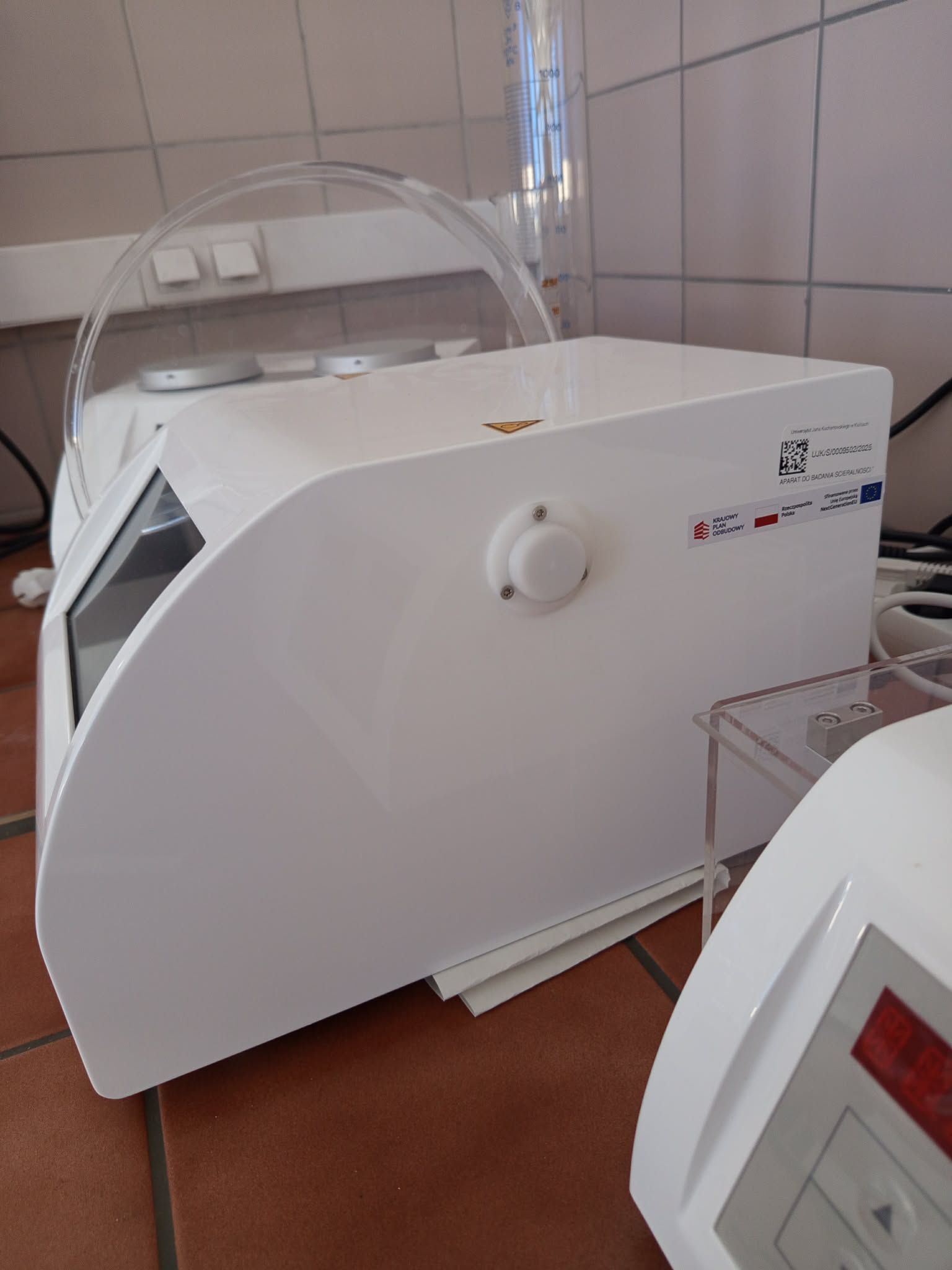 Zdjęcie przedstawia duże białe urządzenie medyczne o zaokrąglonej obudowie, wyposażone w przezroczystą osłonę oraz pokrętło sterujące. Za urządzeniem znajduje się pionowy przezroczysty cylinder pomiarowy z oznaczeniami. Na obudowie widoczne są etykiety z kodem QR oraz informacjami dotyczącymi urządzenia i finansowania.