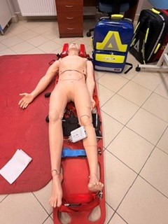 Zdjęcie przedstawia manekin szkoleniowy ułożony na czerwonej macie w sali dydaktycznej. Wokół manekina znajdują się pasy, sprzęt oraz torby medyczne i ratownicze, w tym torba z dużym żółtym symbolem. W tle widoczne są płytki podłogowe oraz drewniana szafka.