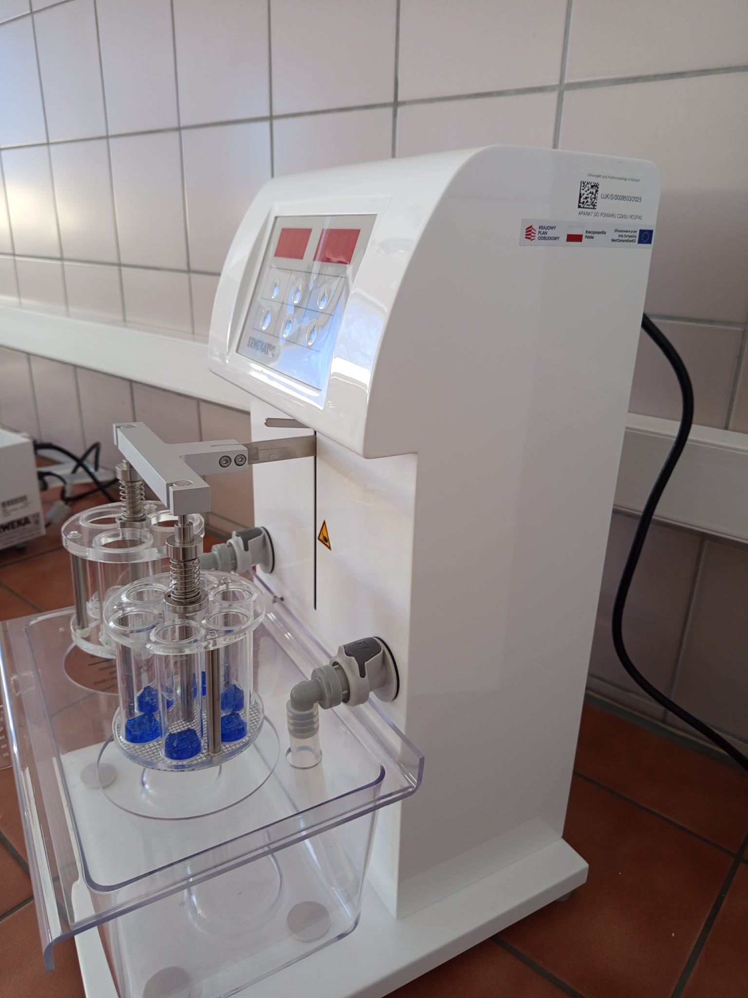 Zdjęcie przedstawia laboratoryjne urządzenie stołowe z panelem sterowania, wyświetlaczem oraz przyciskami. Z przodu znajduje się przezroczysty uchwyt z cylindrycznymi probówkami, połączony z urządzeniem za pomocą prętów, sprężyn, przewodów i zacisków. Na obudowie widoczne są oznaczenia, logotypy oraz kod QR, a dolny mechanizm osłania przezroczysta obudowa.