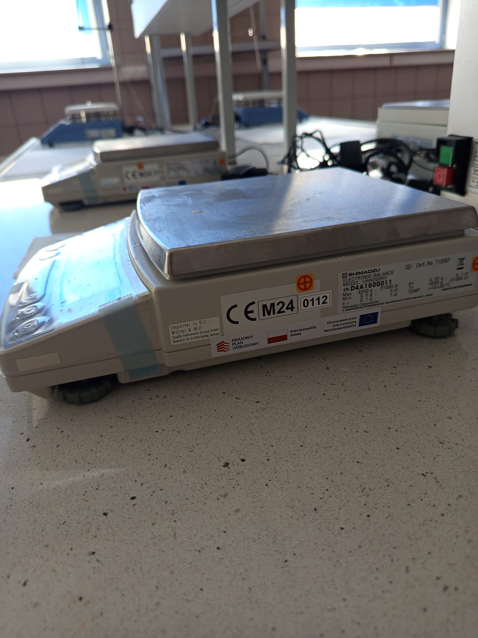Zdjęcie przedstawia laboratoryjną wagę elektroniczną Shimadzu z dużą platformą ze stali nierdzewnej oraz przezroczystą osłoną przeciwwiatrową. Na froncie widoczne są oznaczenia modelu, symbole ostrzegawcze oraz etykiety dotyczące finansowania ze środków polskich i Unii Europejskiej. W tle znajdują się dodatkowe urządzenia laboratoryjne, przewody oraz okna zapewniające dostęp do światła dziennego.
