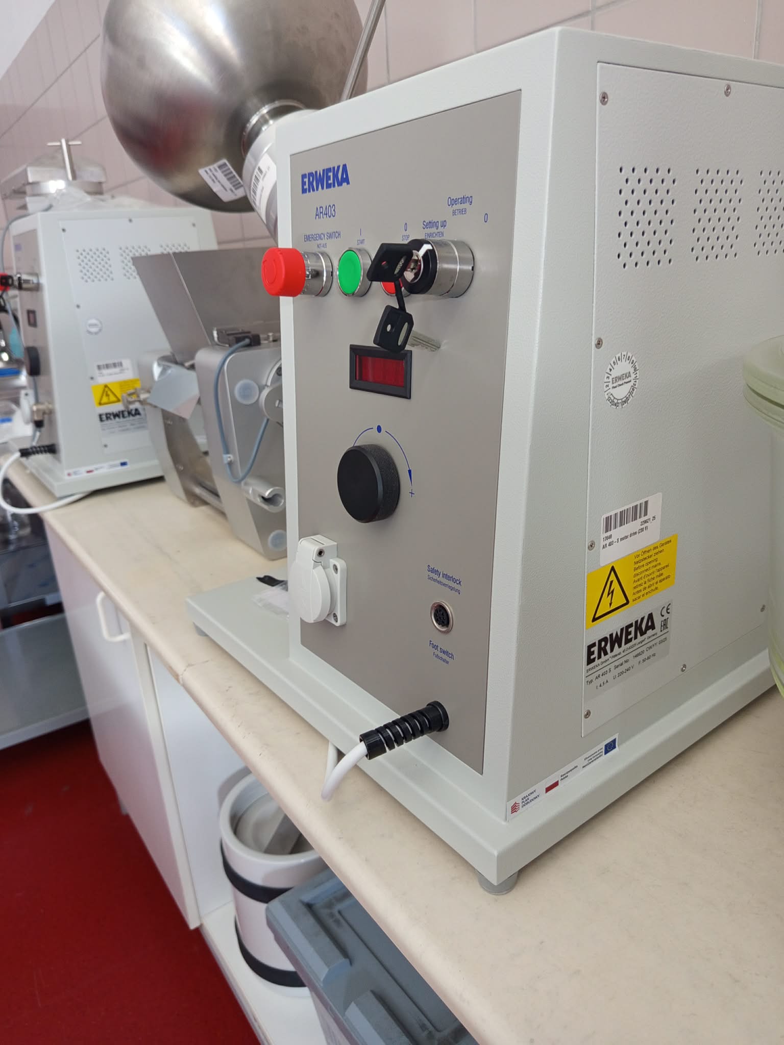 Zdjęcie przedstawia dużą maszynę laboratoryjną ERWEKA AR403 z panelem sterowania oraz metalowym zbiornikiem umieszczonym w górnej części urządzenia. Panel wyposażony jest w przyciski, wyłącznik awaryjny, przełącznik kluczykowy oraz pokrętło regulacyjne. Na bocznej części widoczna jest kratka wentylacyjna i oznaczenie ostrzegawcze, a w tle znajdują się dodatkowe urządzenia laboratoryjne.