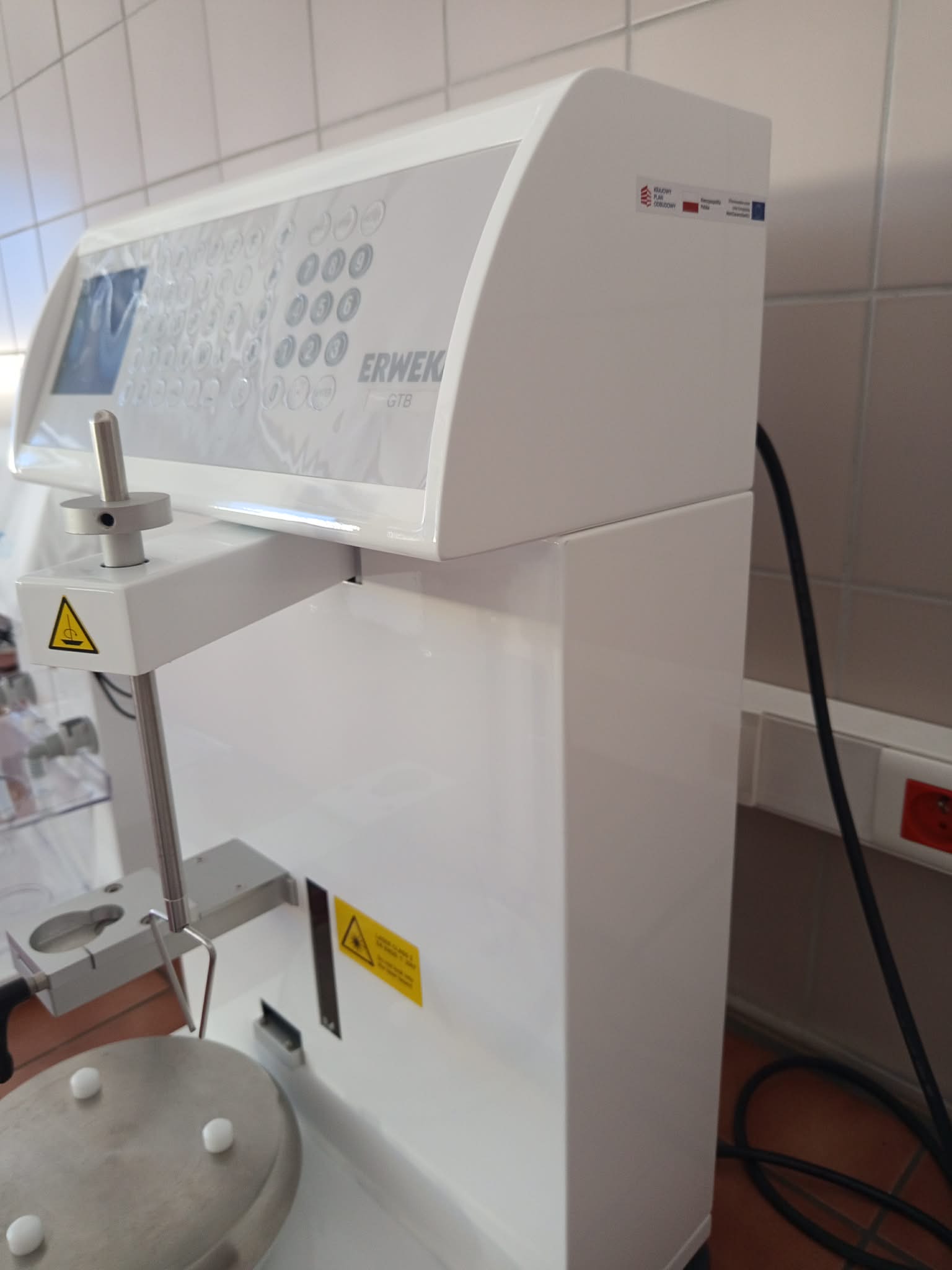 Zdjęcie przedstawia duże urządzenie laboratoryjne ERNEK GTB z panelem sterowania wyposażonym w wyświetlacz i rzędy przycisków. Z przodu znajdują się metalowy pręt regulacyjny oraz okrągła platforma robocza, a na obudowie widoczne są ostrzegawcze oznaczenia bezpieczeństwa. Do urządzenia podłączone są przewody, a w tle znajduje się dodatkowe wyposażenie laboratoryjne.