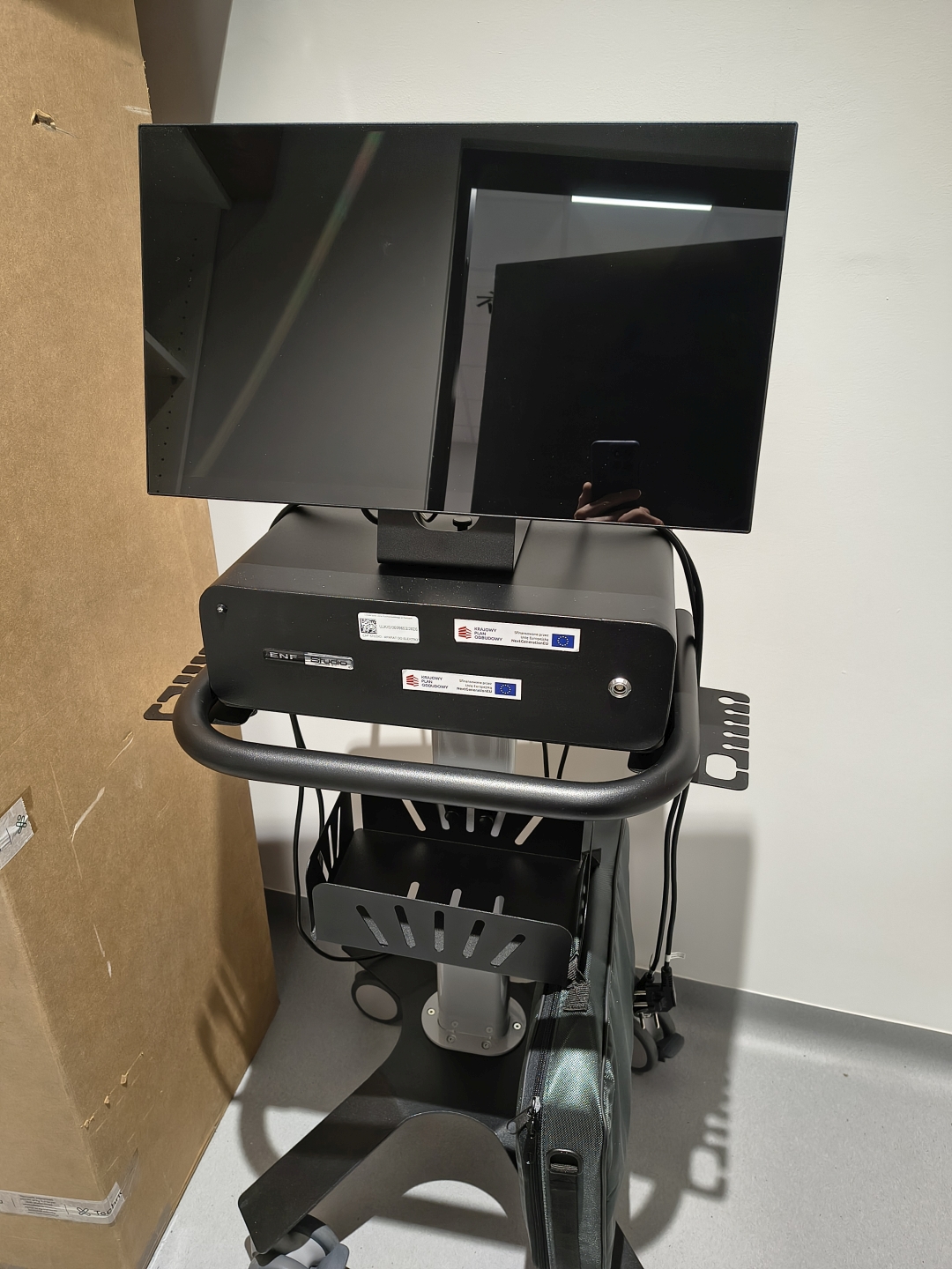 Zdjęcie przedstawia czarny wózek ze sprzętem medycznym wyposażony w monitor oraz panel sterowania. Na wózku znajdują się otwarte kosze na wyposażenie, przewody i elementy mocujące, a na urządzeniu widoczne jest oznaczenie „ENF DUCO”. W tle znajduje się kartonowy panel, biała ściana oraz szara podłoga.