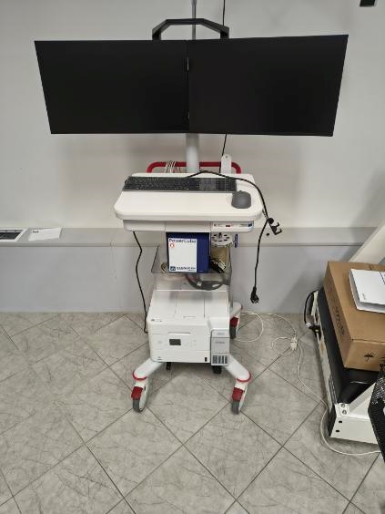 Zdjęcie przedstawia mobilny wózek ze sprzętem pomiarowym wyposażony w dwa duże monitory oraz konsolę sterującą z klawiaturą i pokrętłem. W dolnej części znajdują się przewody oraz kółka umożliwiające przemieszczanie urządzenia. W tle widoczne są dodatkowe wyposażenie oraz przewody znajdujące się na podłodze.