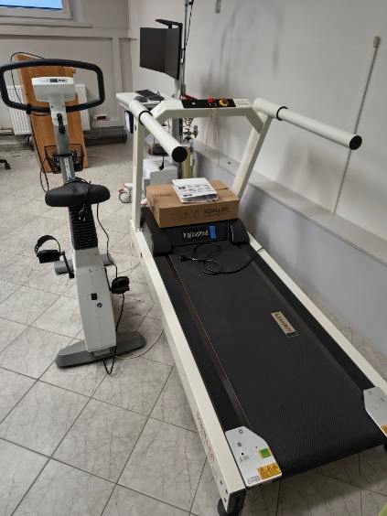Zdjęcie przedstawia stanowisko do badań wysiłkowych wyposażone w bieżnię z poręczami, panelem sterowania oraz szerokim pasem biegowym. Obok znajduje się rower stacjonarny z panelem wyświetlacza. Za bieżnią widoczny jest monitor, a sprzęt ustawiono na jasnej, płytkowej podłodze z widocznymi przewodami elektrycznymi.