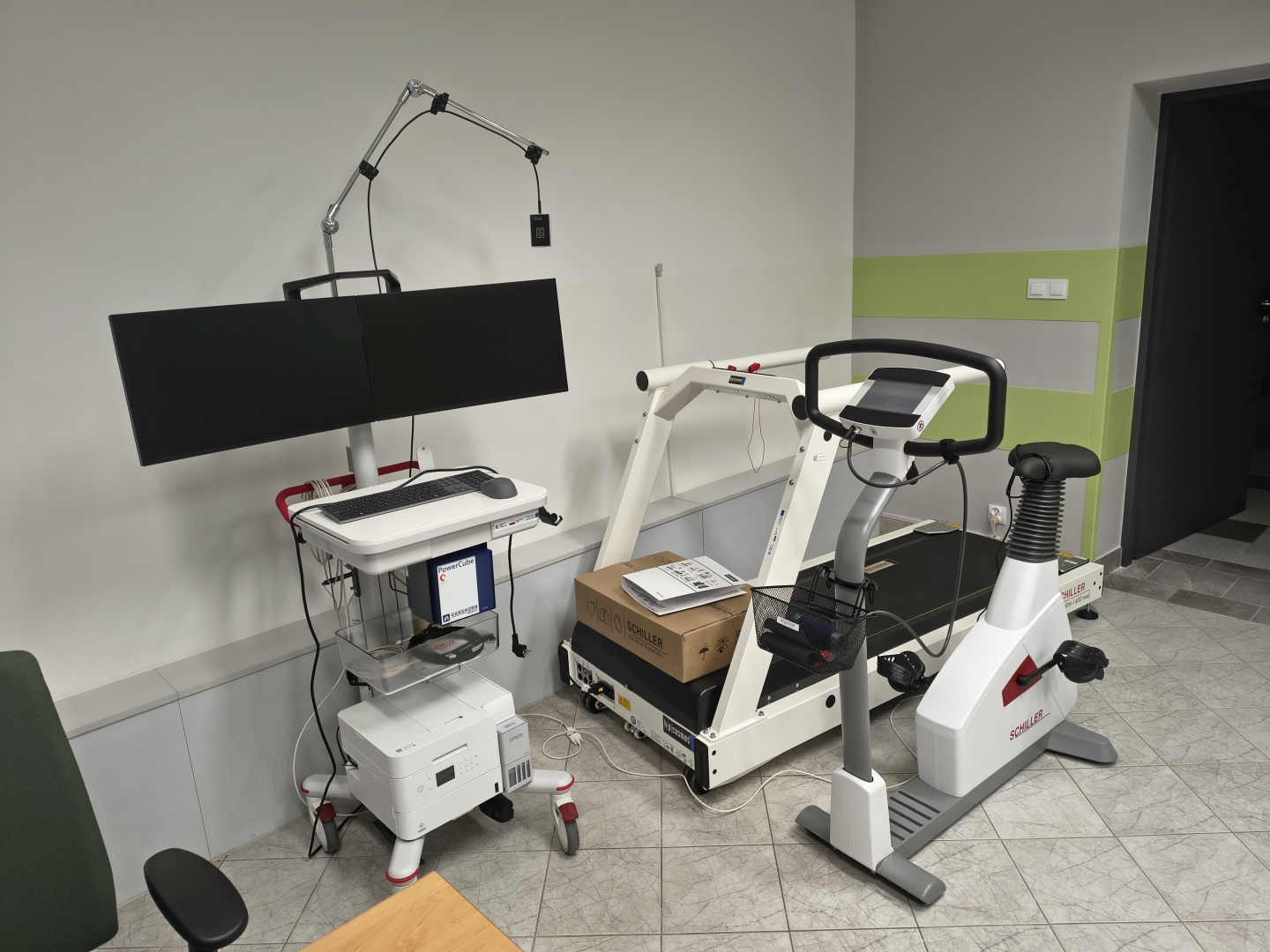 Zdjęcie przedstawia salę do badań wysiłkowych wyposażoną w rower stacjonarny oraz bieżnię. W pomieszczeniu znajdują się również monitory komputerowe, stanowisko komputerowe, drukarka oraz dodatkowe wyposażenie medyczne. W tle widoczne są przewody, wózek medyczny i elementy wyposażenia technicznego.