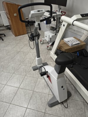 Zdjęcie przedstawia stacjonarny rower treningowy lub rehabilitacyjny wyposażony w regulowane elementy, pedały z paskami oraz przewody. Obok znajduje się duże urządzenie do ćwiczeń z siedziskiem. W tle widoczne są dodatkowe przyrządy treningowe, drzwi oraz grzejnik.