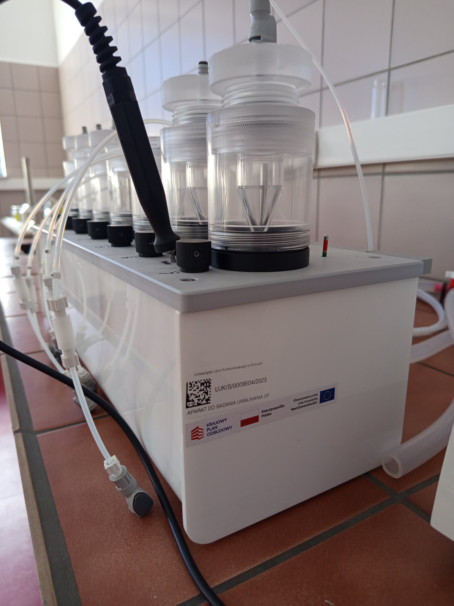 Zdjęcie przedstawia aparaturę laboratoryjną umieszczoną na białej szafce, z kilkoma przezroczystymi cylindrycznymi naczyniami połączonymi przewodami. Z urządzenia wychodzą dodatkowe rurki, złącza oraz gruby przewód. Na froncie szafki widoczna jest etykieta dotycząca finansowania i identyfikacji wyposażenia z kodem QR.
