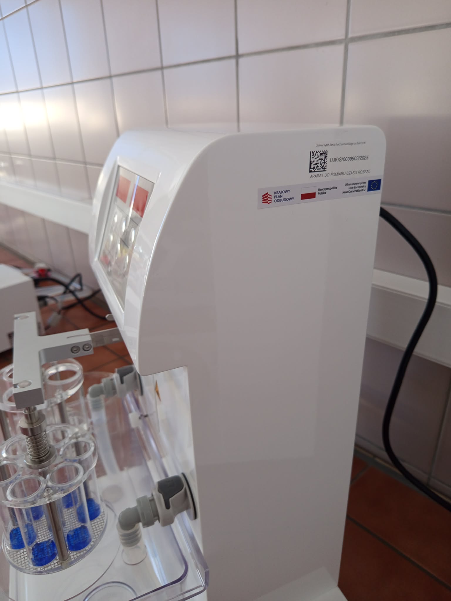 Zdjęcie przedstawia duże białe urządzenie laboratoryjne z zaokrągloną obudową oraz częściowo widocznym panelem sterowania. W dolnej części znajdują się przezroczyste uchwyty z niebieskimi elementami lub cieczą oraz przewody i złącza. Na obudowie widoczna jest etykieta z kodem QR i polskim opisem.