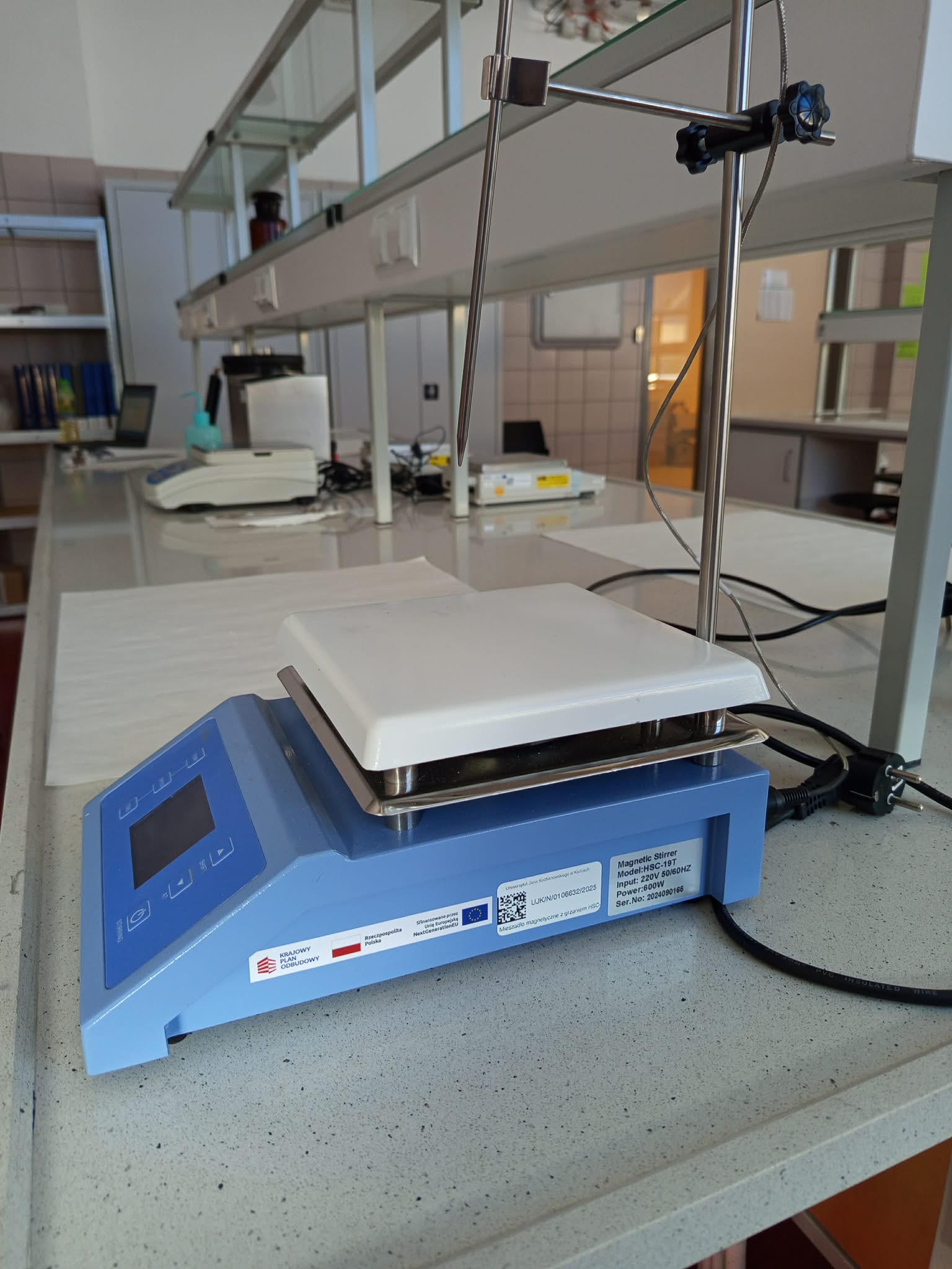 Zdjęcie przedstawia niebieskie mieszadło magnetyczne z funkcją podgrzewania, wyposażone w płytę grzejną, panel sterowania z wyświetlaczem oraz przewód zasilający. Obok urządzenia znajduje się statyw z zaciskami i metalowym prętem lub sondą umieszczoną nad płytą. W tle widoczne są stanowiska laboratoryjne, półki, naczynia oraz dodatkowe wyposażenie.